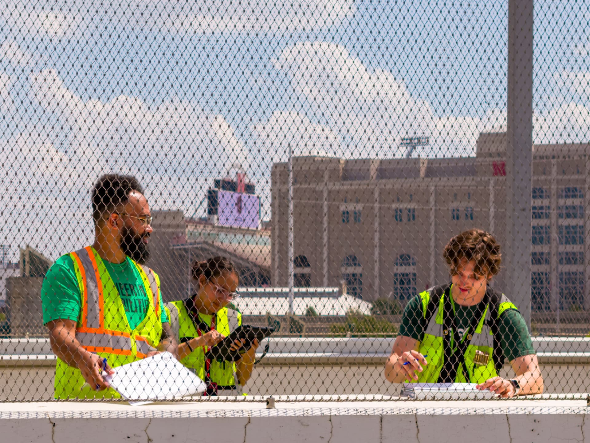Workers in safety vests review plans outdoors near a fence, with a building in the background.