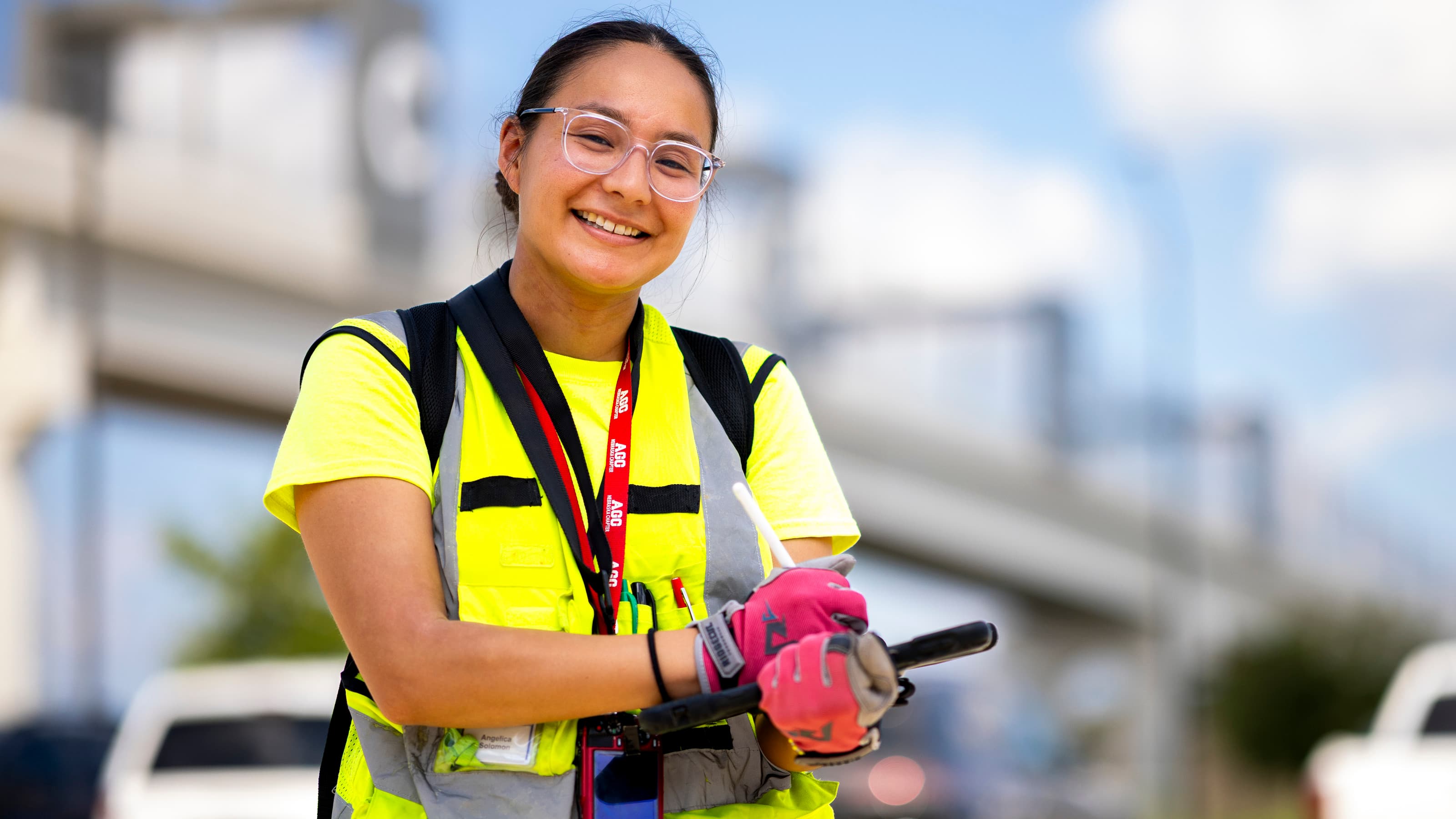 Smiling worker wearing a safety vest, gloves, and glasses, standing outdoors near a roadway.