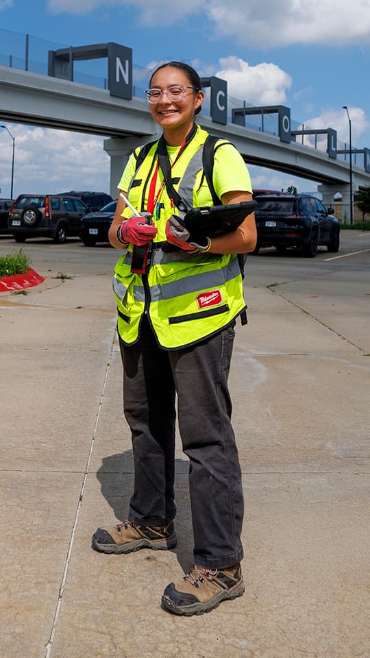 A smiling worker in a safety vest holds a tablet and radio in a parking lot under a sunny sky.