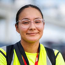 Woman with glasses smiling, wearing a bright yellow vest and lanyard outdoors.