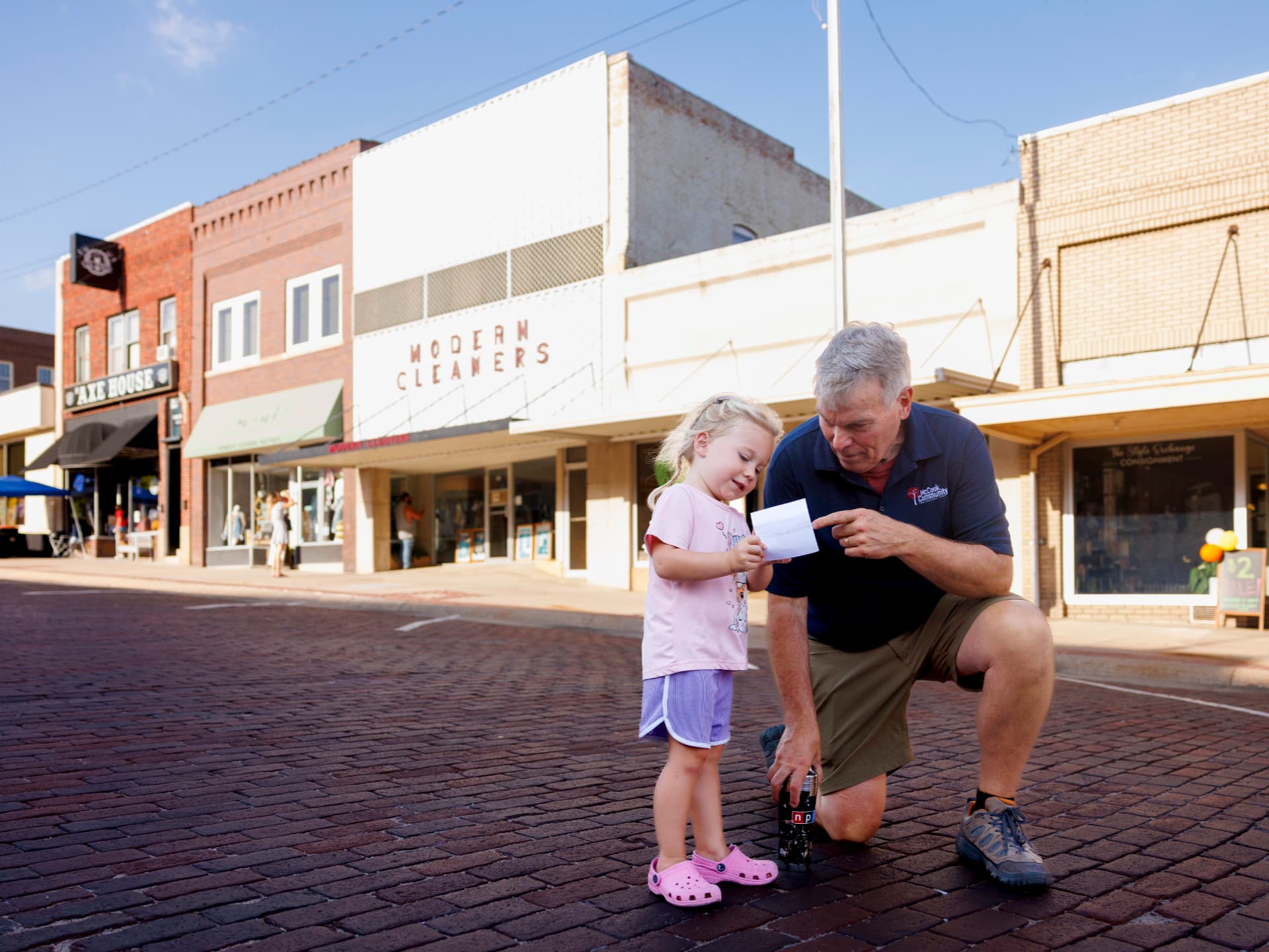 Man kneeling with a young girl, holding paper on a brick street, storefronts in the background.