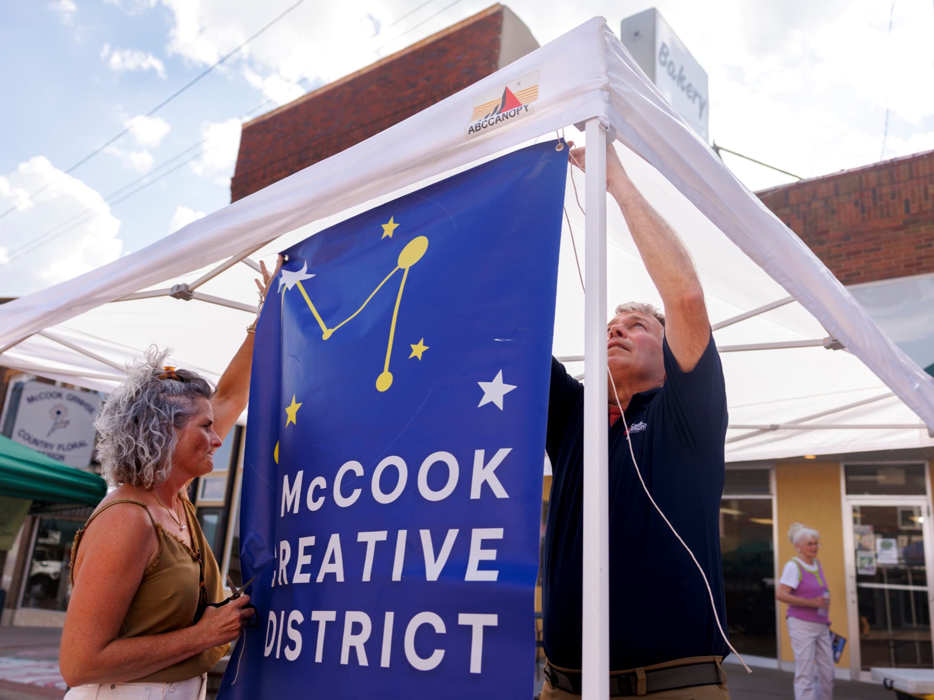 Two people set up a McCook Creative District banner under a canopy outdoors.