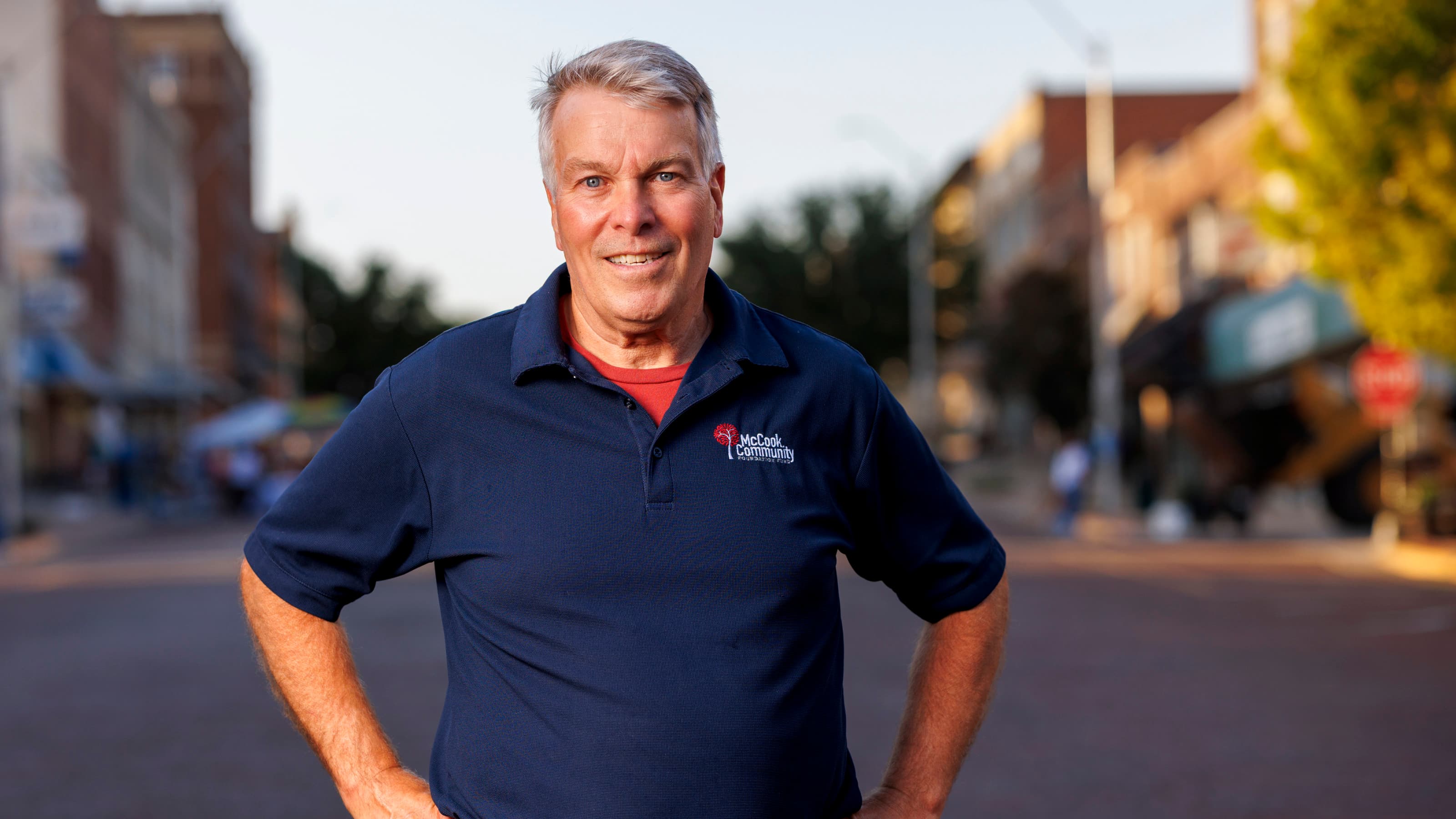 Man smiling, wearing blue polo, standing on a sunlit urban street with blurred buildings and trees.