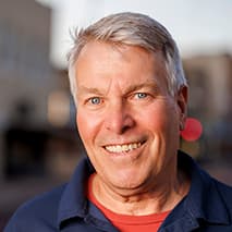 Smiling man wearing a blue shirt outdoors with blurred buildings in the background.