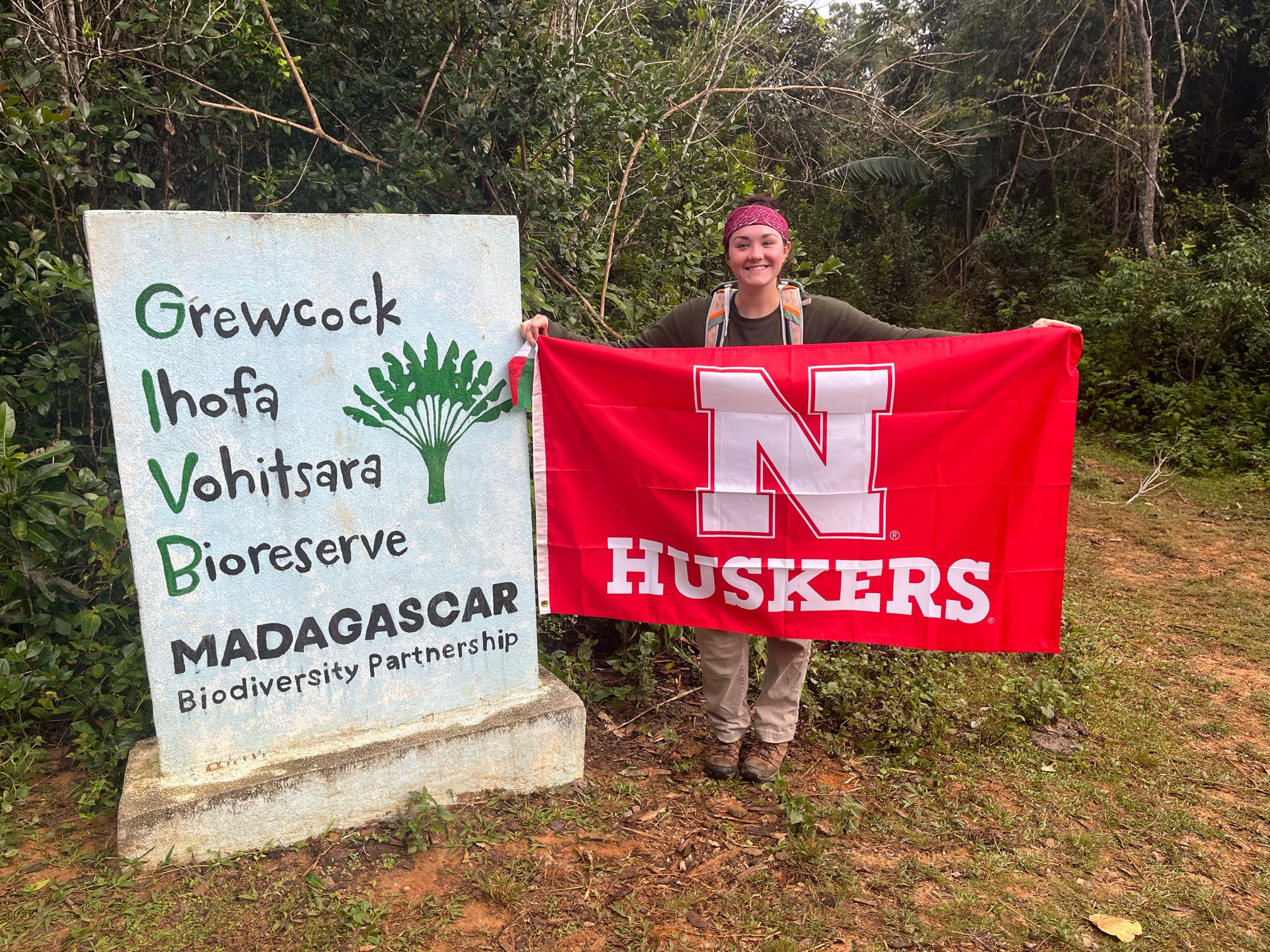 Person holding a red Nebraska Huskers flag next to a biodiversity reserve sign in a forested area.