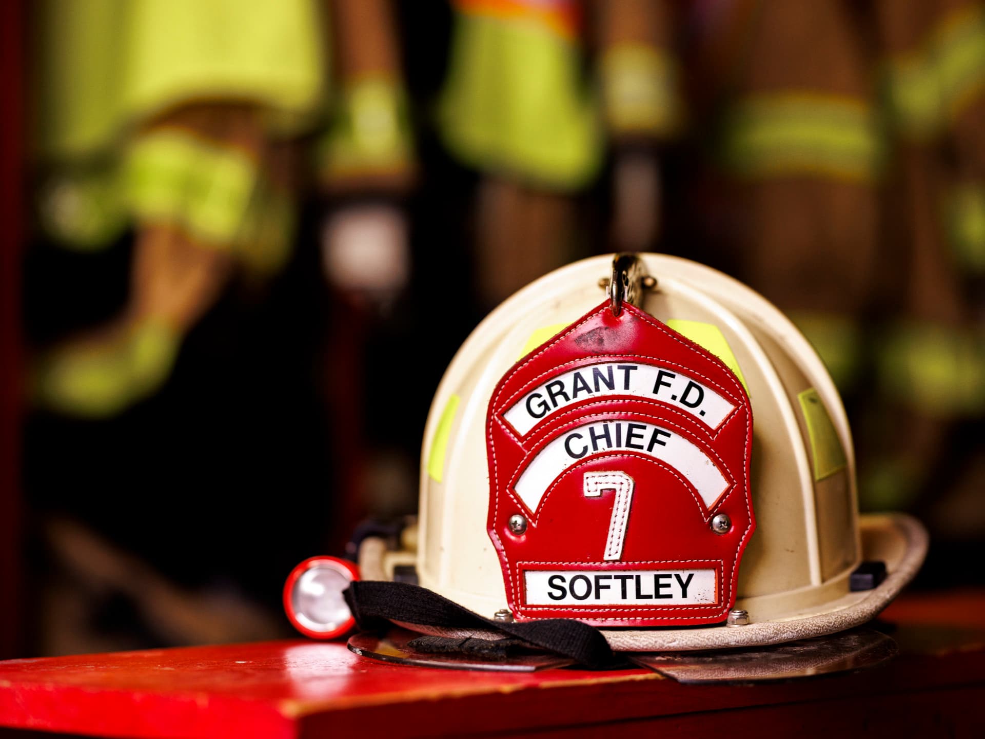 Fire chief helmet with red plaque on a table, blurred firefighting gear in the background.