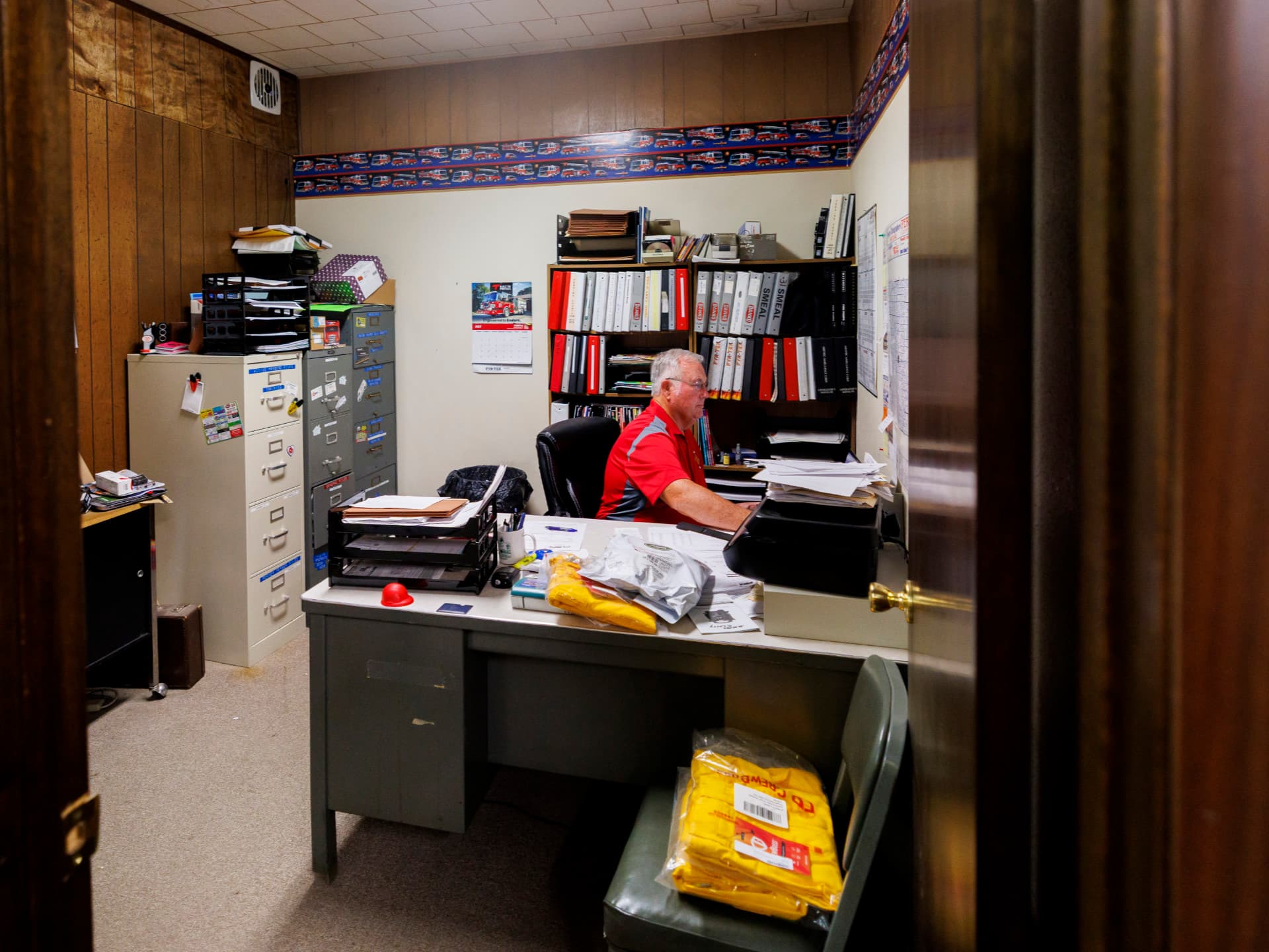 Man in a small office with wooden panels, working at a desk surrounded by files and folders.