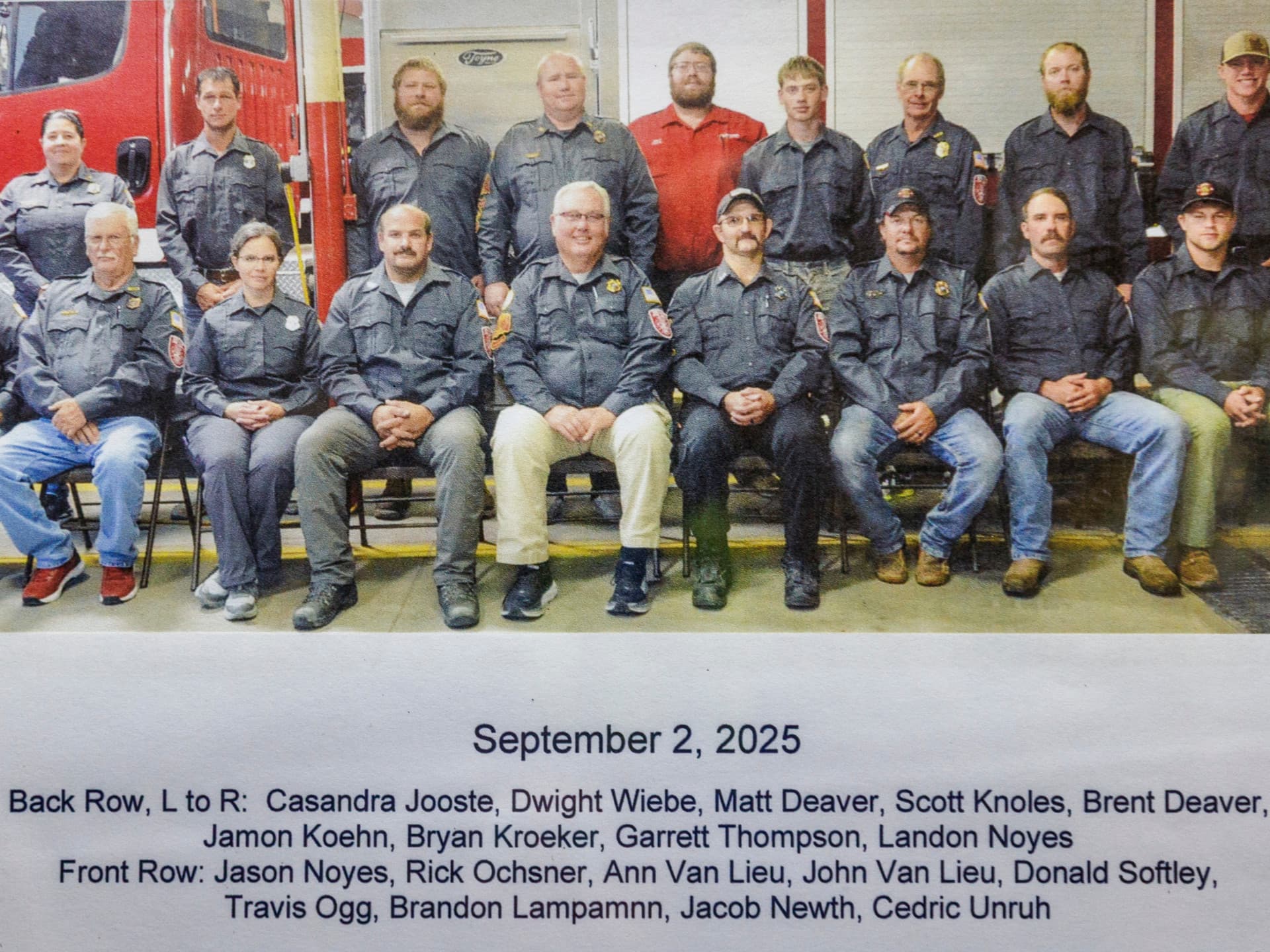 Group of people in uniform posing indoors with red emergency vehicle visible in the background.