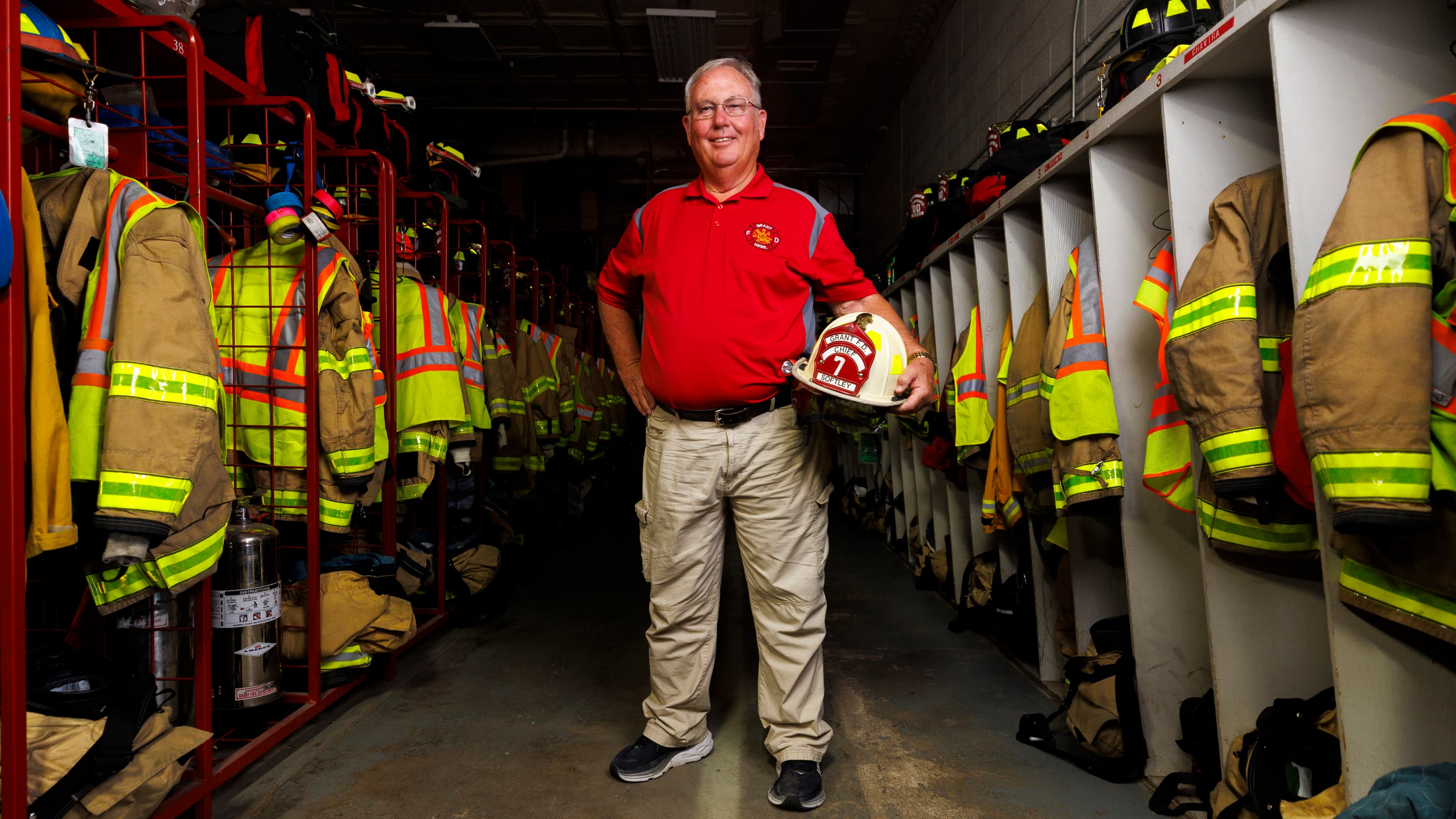 Man in red shirt holding fire helmet, standing in a fire station surrounded by hanging gear.