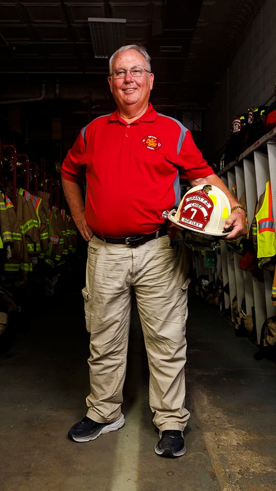 Man in red fire department polo holding fire helmet, standing in a room lined with firefighter gear.
