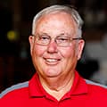 Smiling man wearing glasses and a red polo shirt against a blurred background.