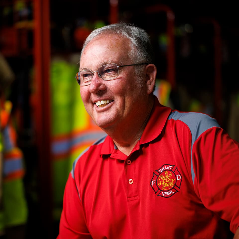 Smiling man in a red fire department shirt, seated in a room with safety gear in the background.