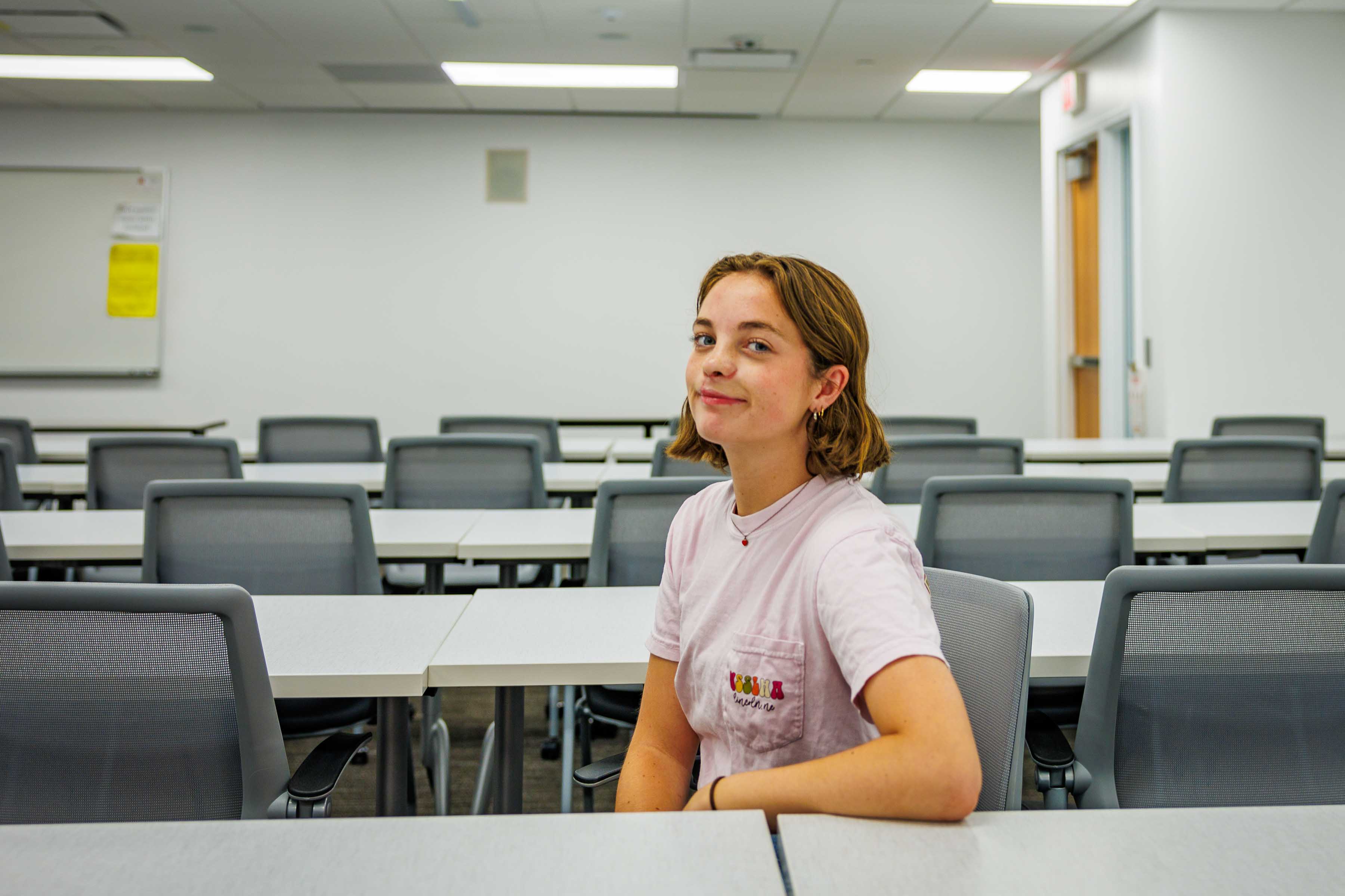 Seated woman smiling in a bright, empty classroom with rows of desks and chairs.