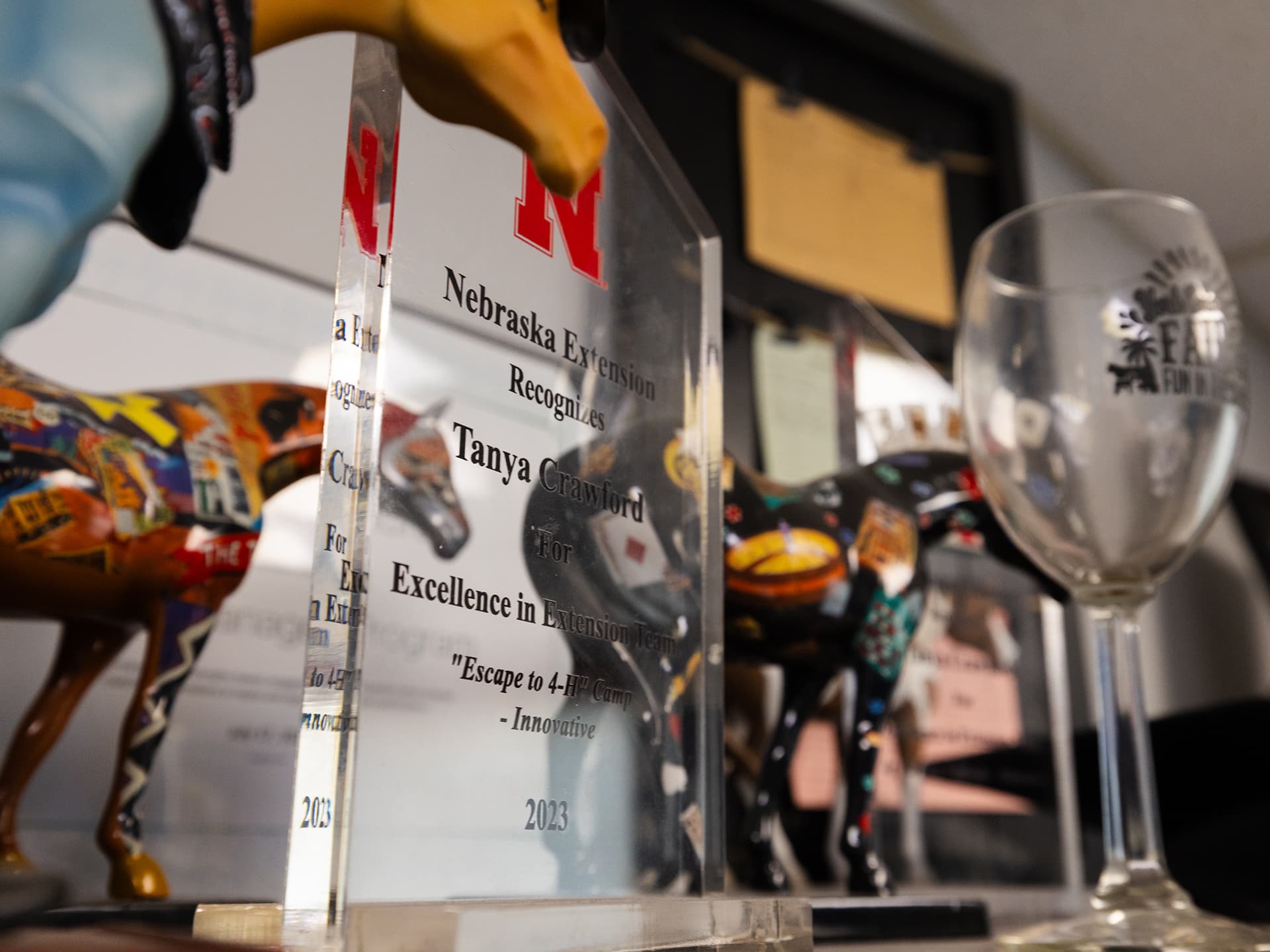 Award plaques, colorful horse figurines, and a wine glass displayed on a shelf.
