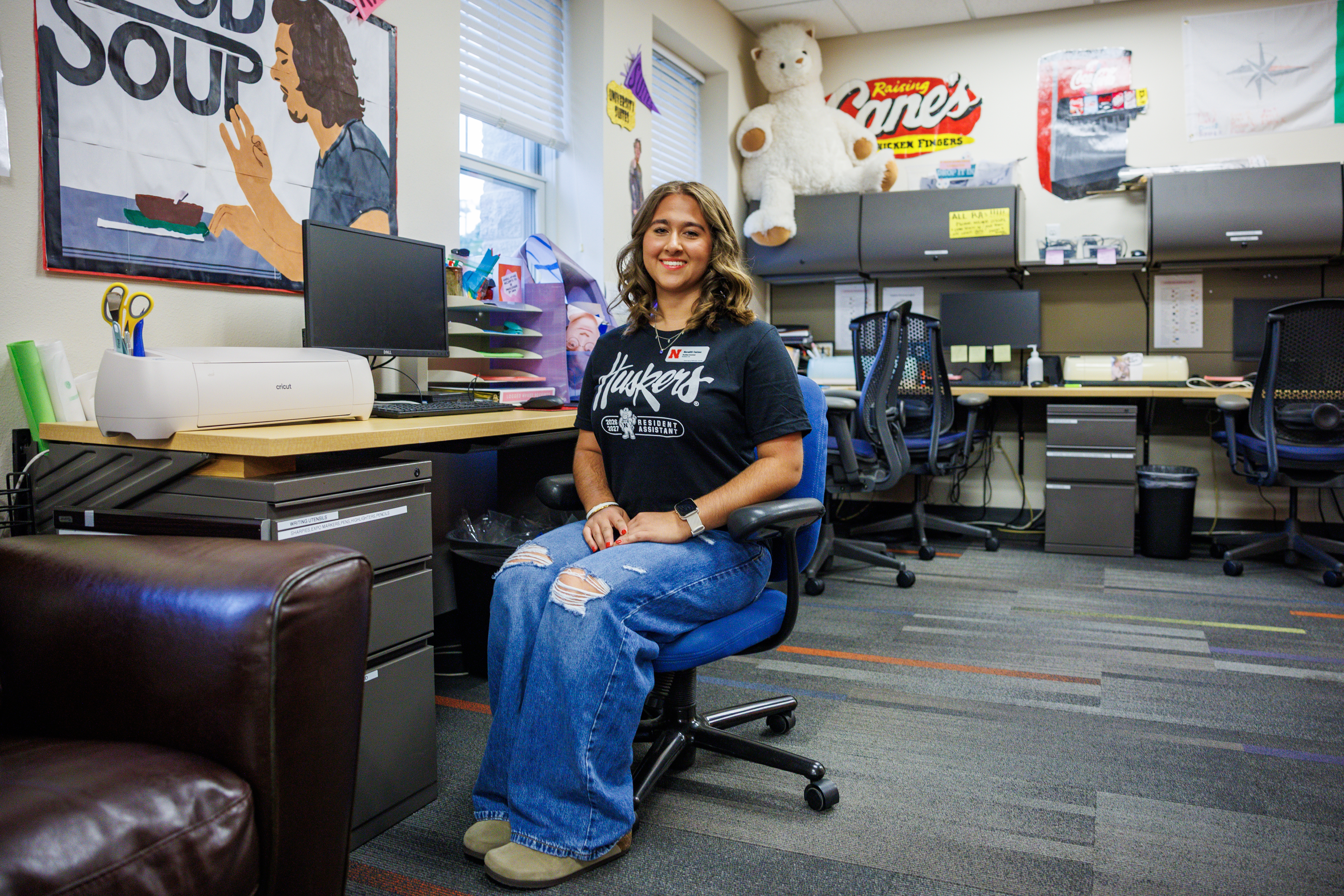 A woman sitting on a chair in a colorful office surrounded by posters, desks, and a large teddy bear.