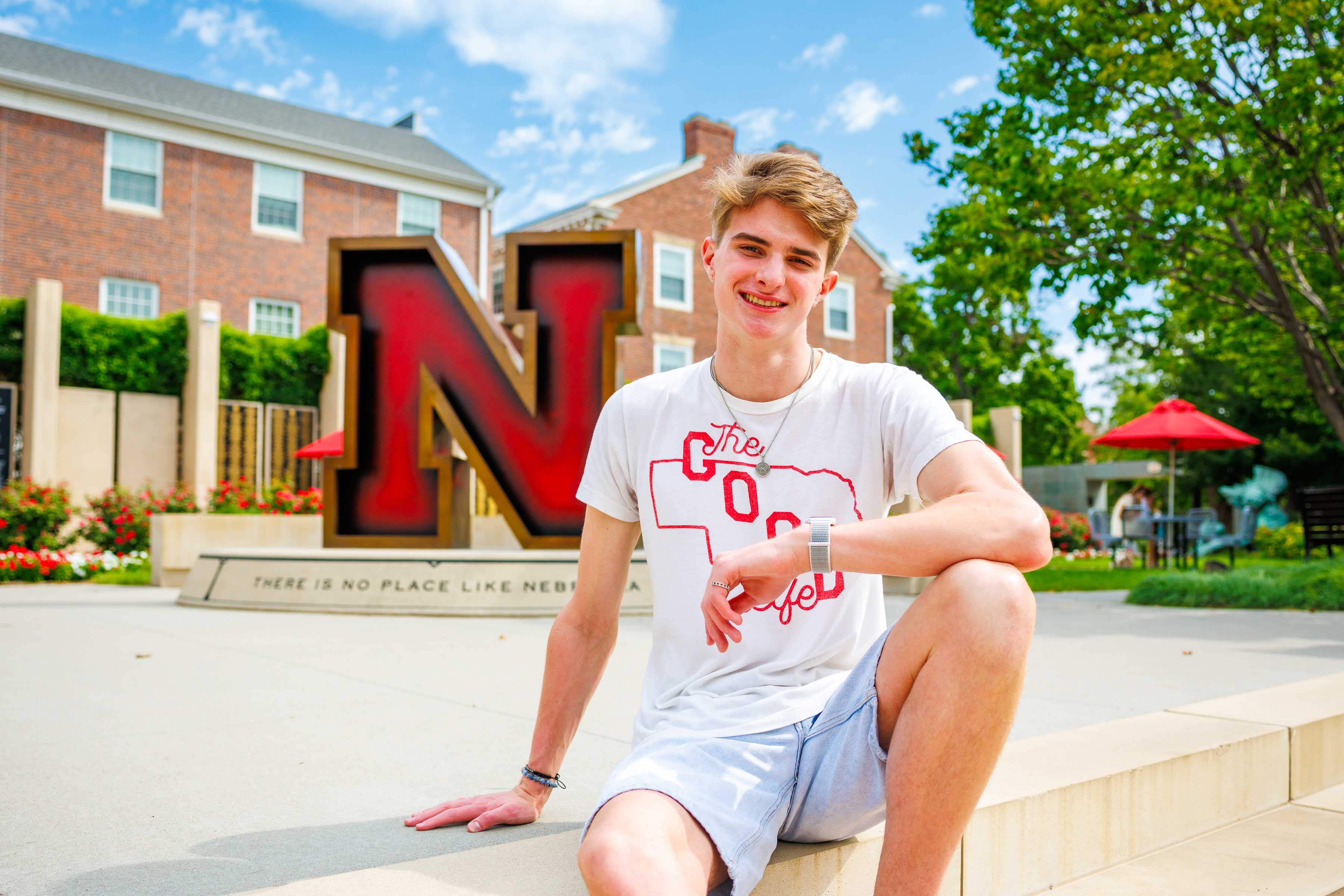 Smiling young man in a white shirt sits outside near a large red "N" sculpture on a sunny day.
