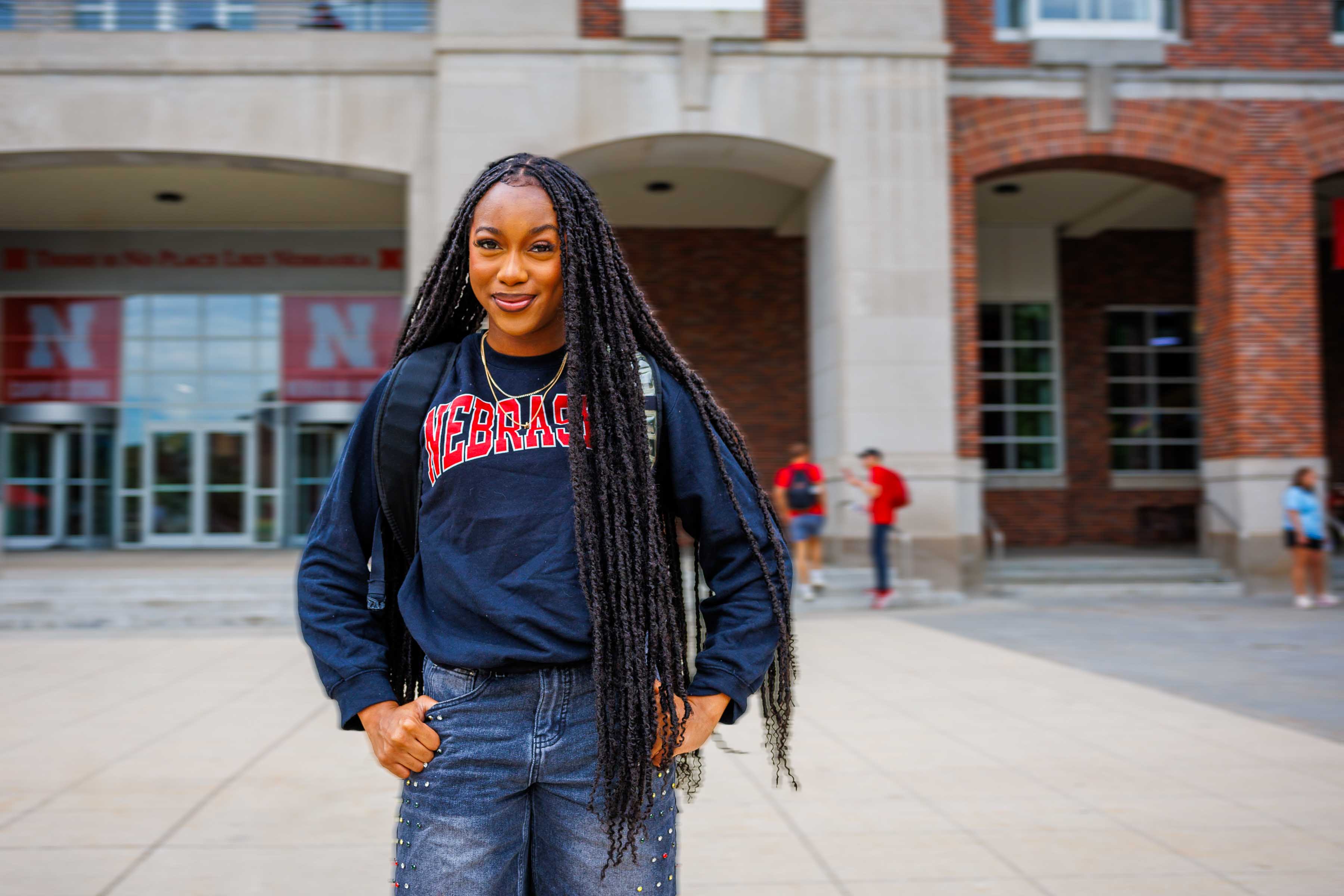 Woman standing outside a building wearing a "Nebraska" sweatshirt, smiling confidently.