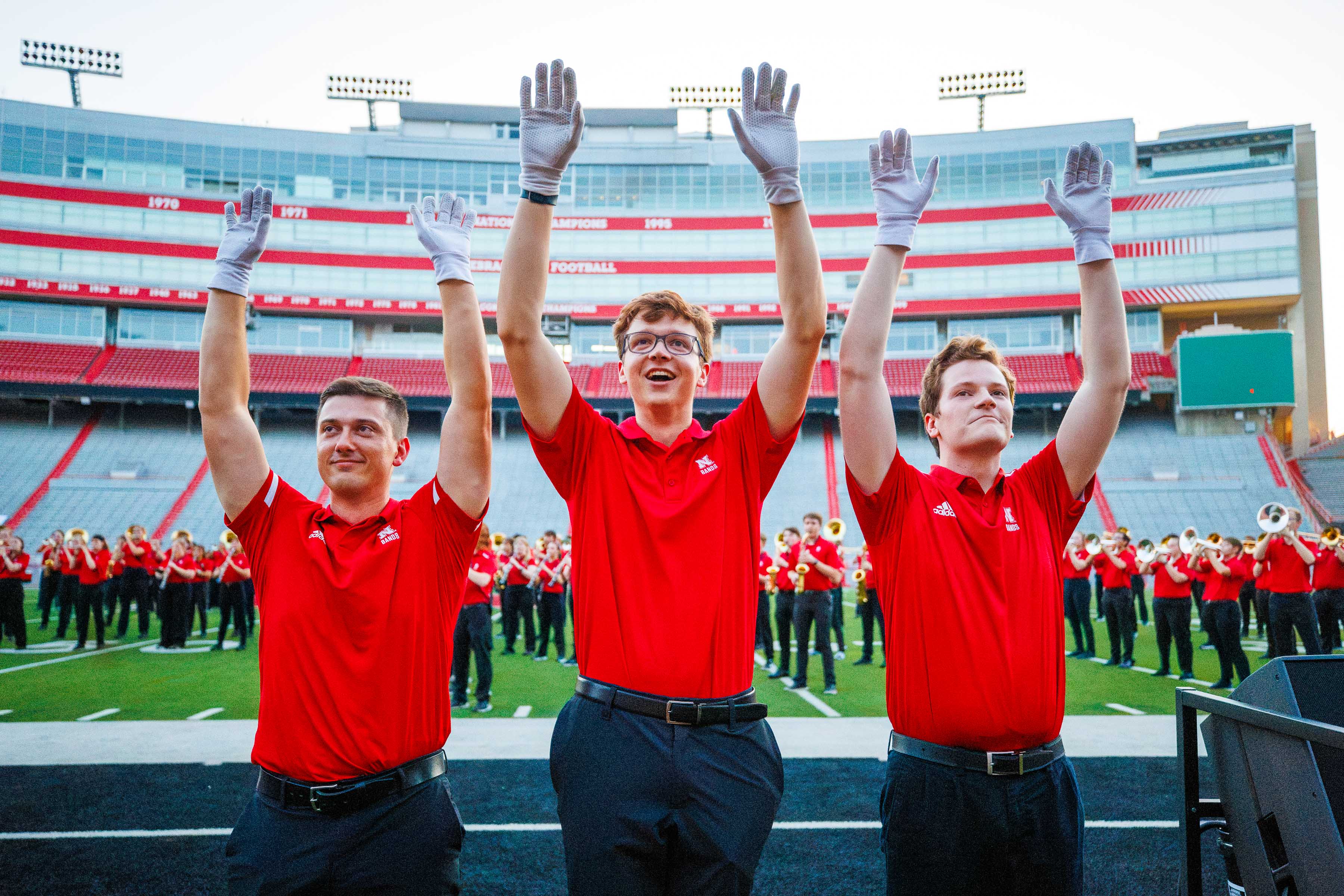 Three drum majors in red shirts raise their arms on a football field with marching band behind them.