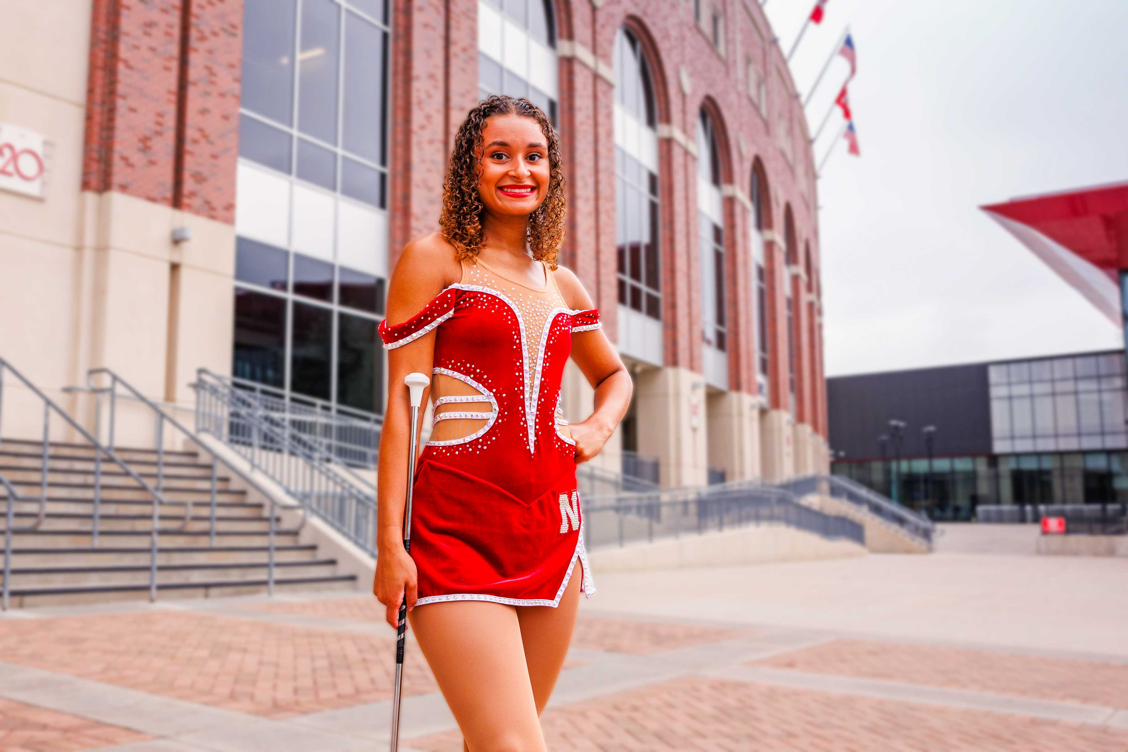 Woman in red baton twirler uniform standing outside near a brick building with stairs and flags.