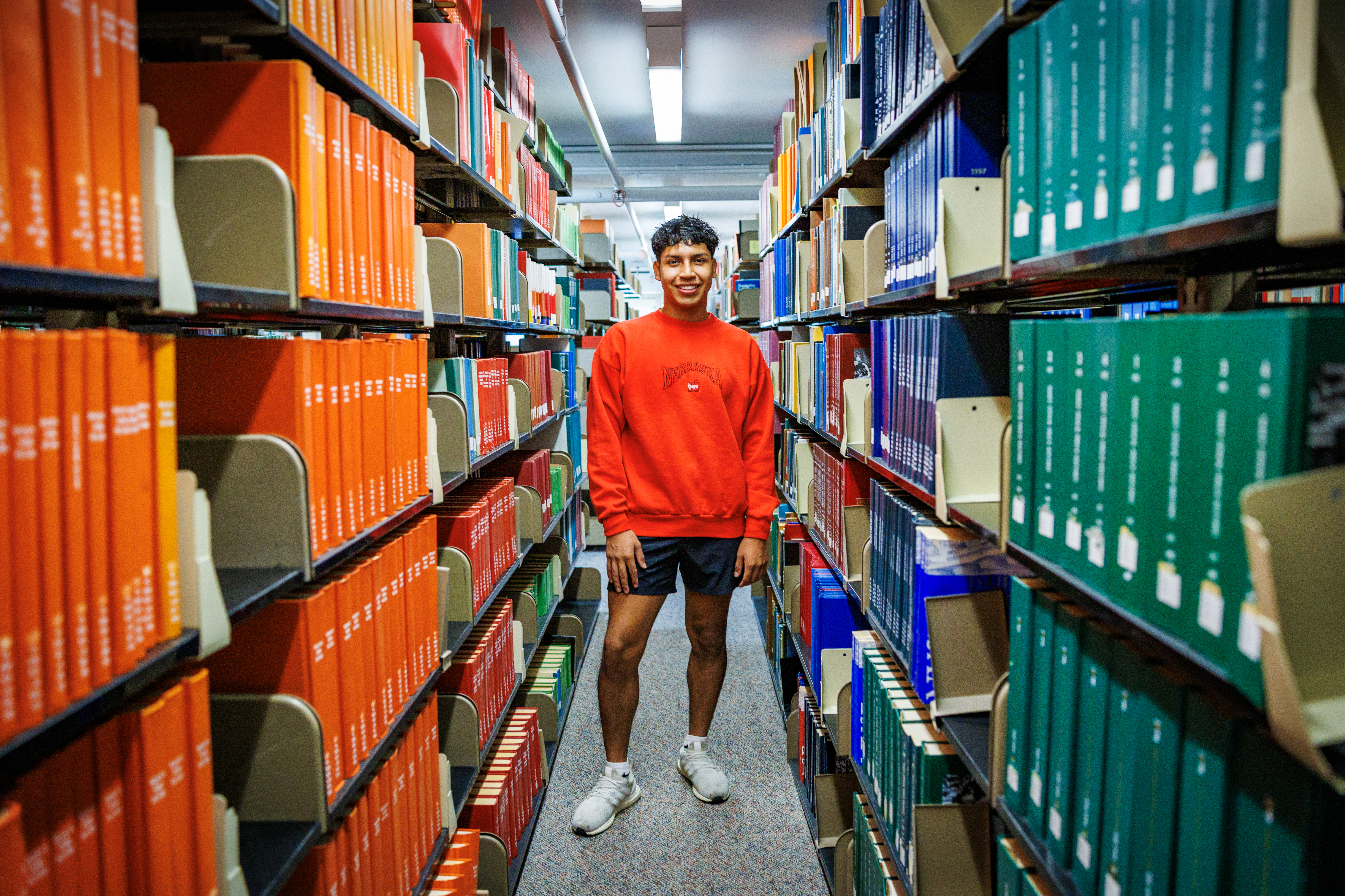 Person in a bright red sweater standing between colorful book-filled library shelves.
