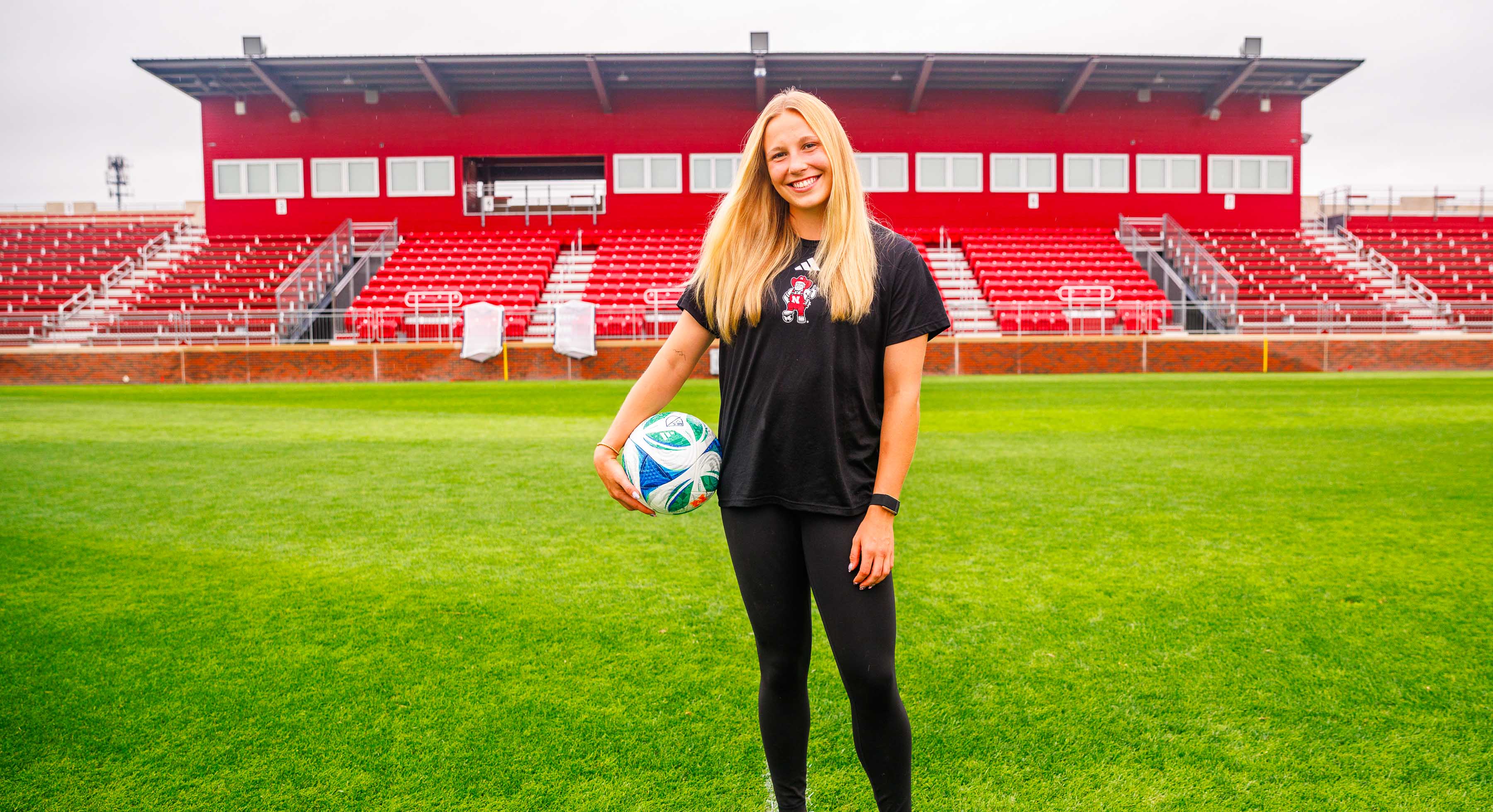 Smiling woman holding a soccer ball on a green field with a red stadium in the background.
