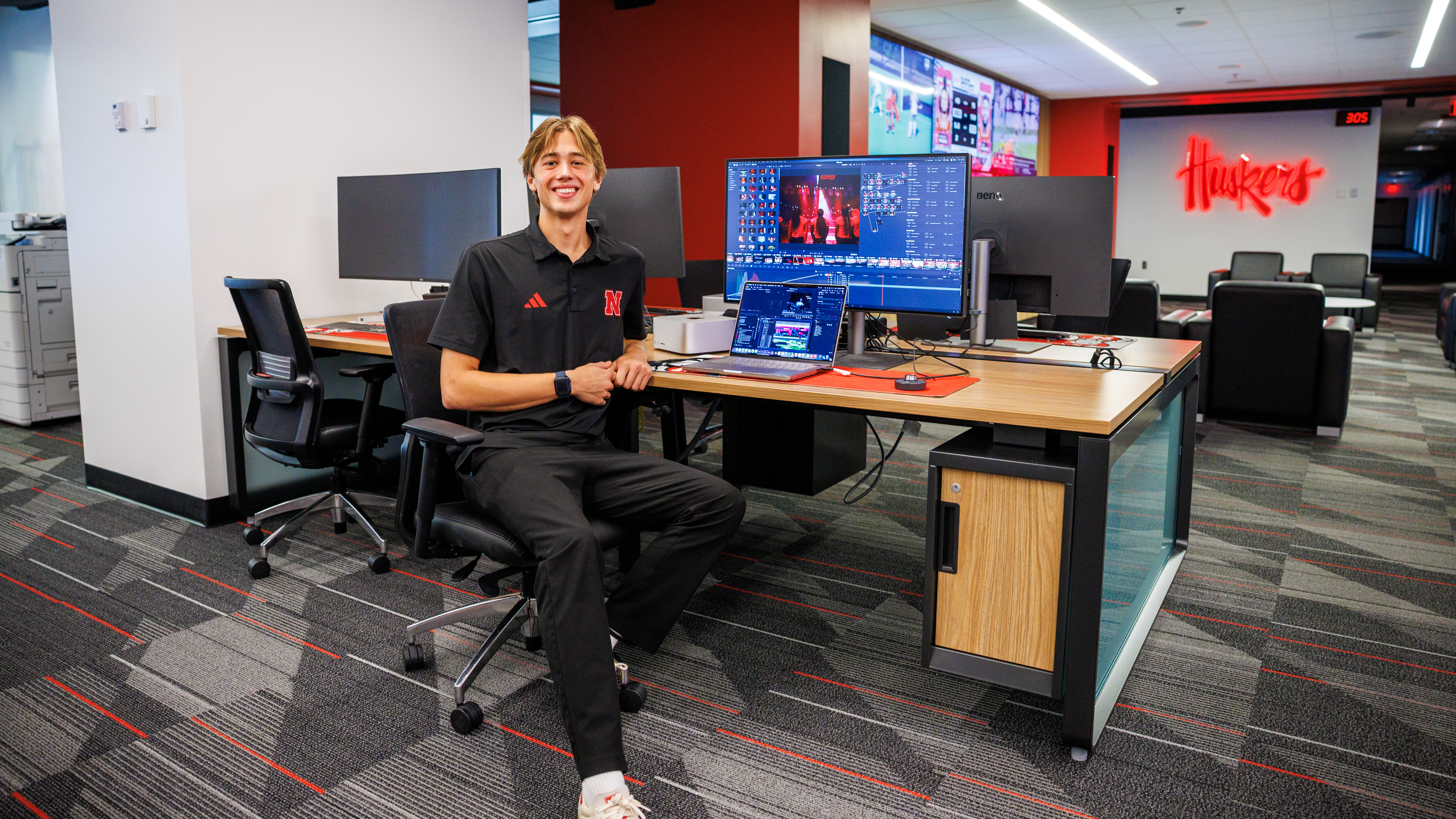 Smiling man seated at a desk in a modern office with computer screens displaying editing software.