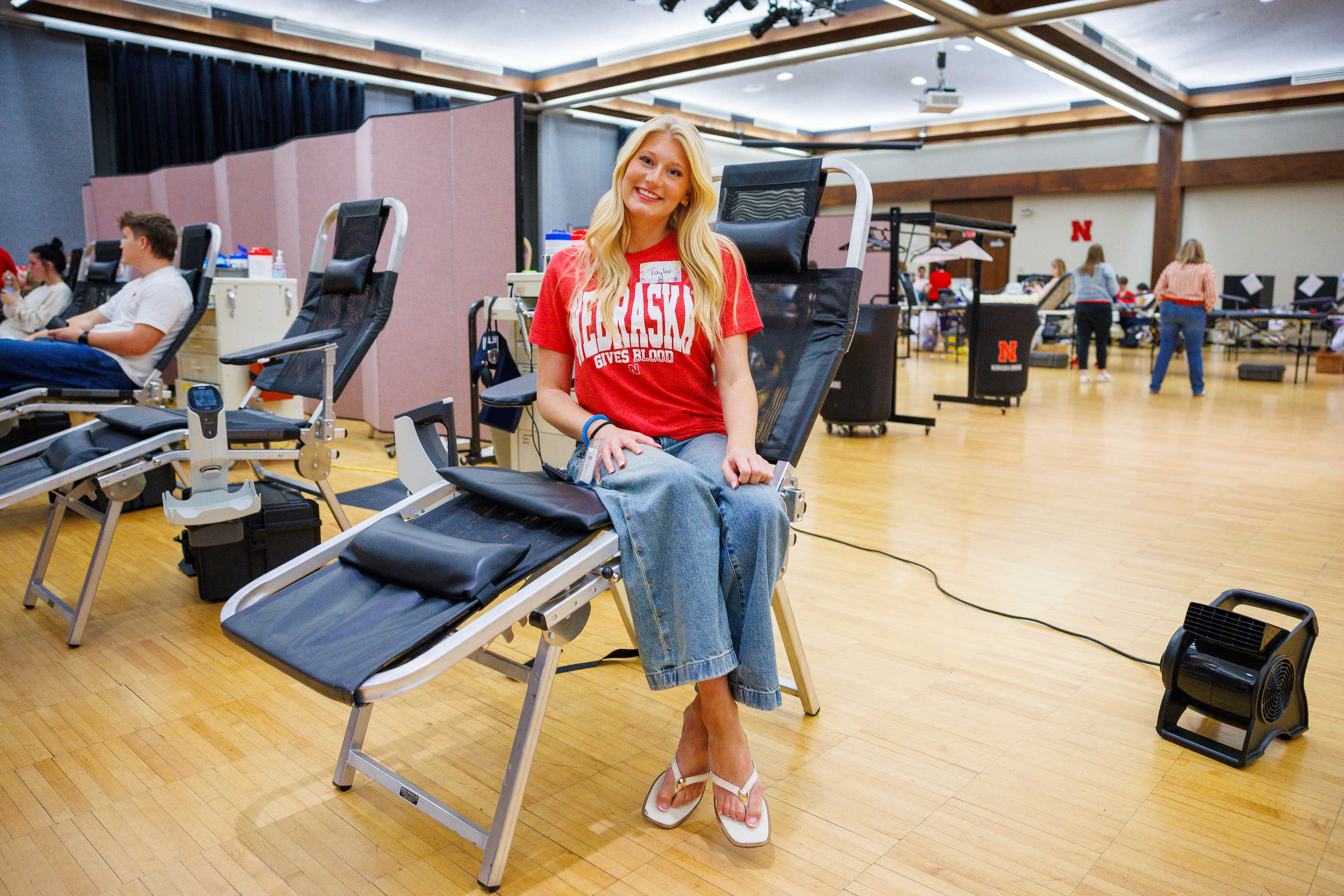 Smiling woman in a Nebraska shirt sits on a blood donation chair in a bright gym.