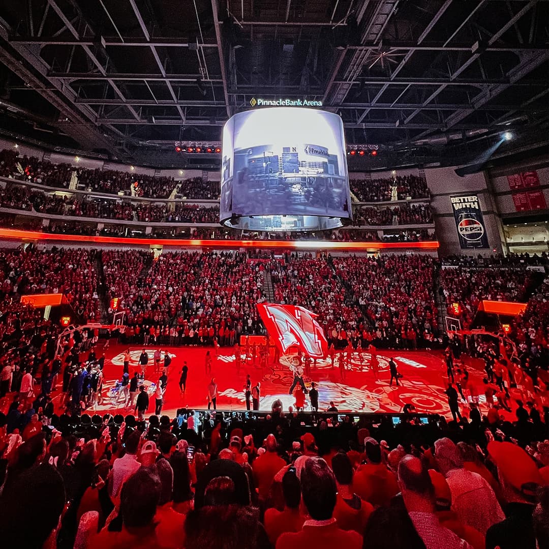 Herbie Husker waves a large Nebraska 'N' flag on a basketball court bathed in red lights at Pinnacle Bank Arena.
