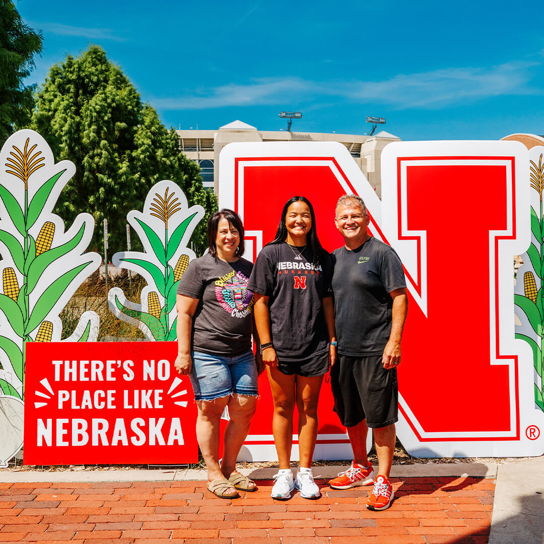 Three people pose smiling near a large red 'N' and greenery under a bright blue sky during Move-In.