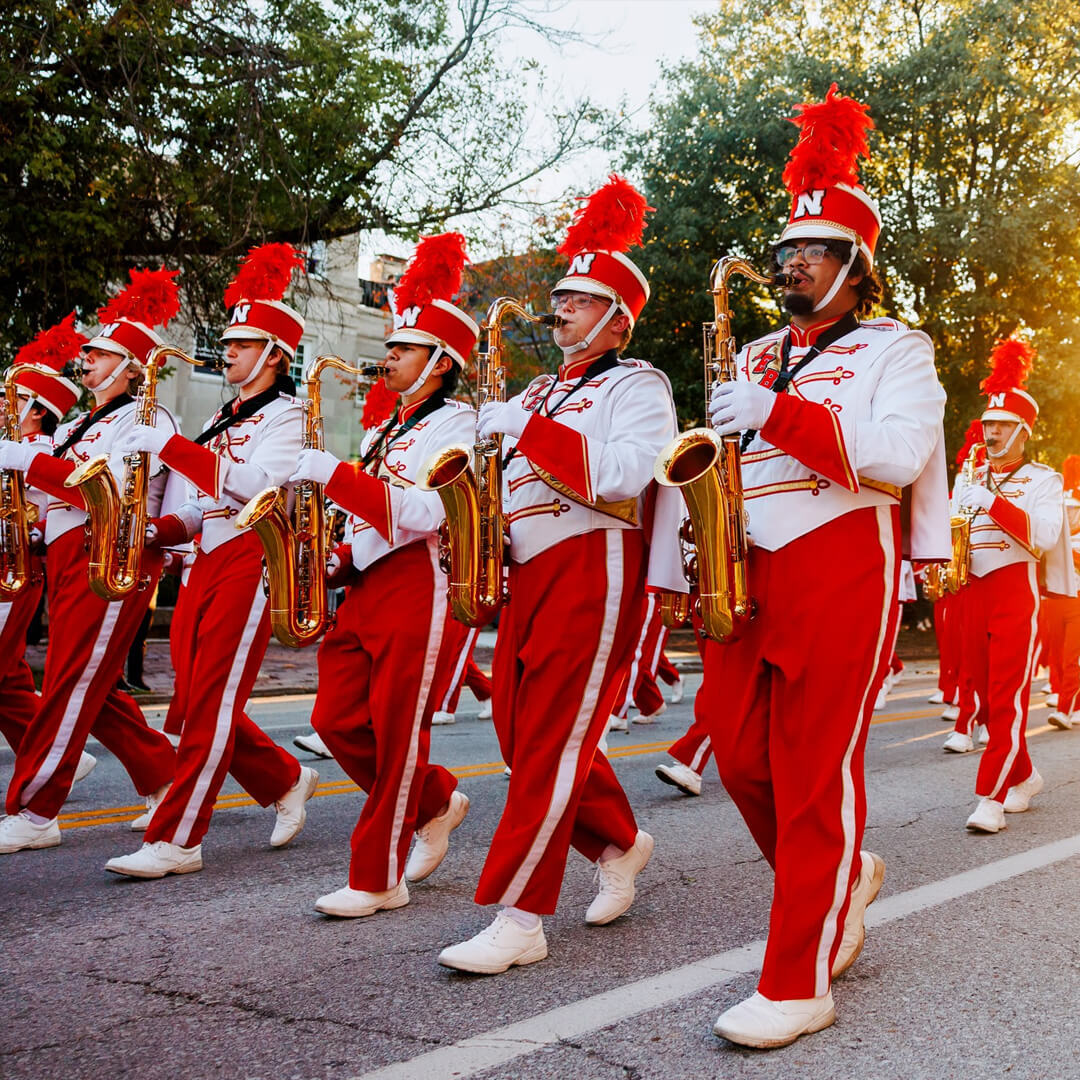 Cornhusker Marching Band saxophone players in red uniforms march in Homecoming Parade on a sunny day.
