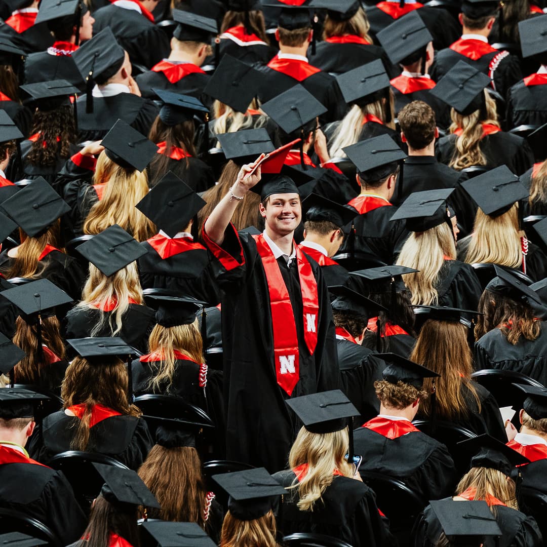 Graduates in black caps and gowns with red accents gathered at a commencement.