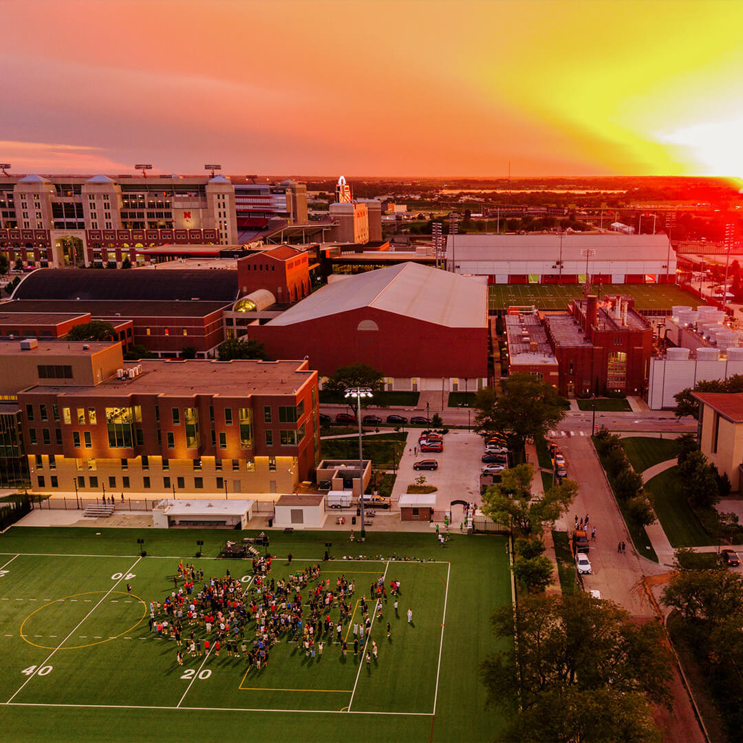 Group of students gather on Mabel Lee Fields during Playfair event at sunset, surrounded by buildings and vibrant orange sky.