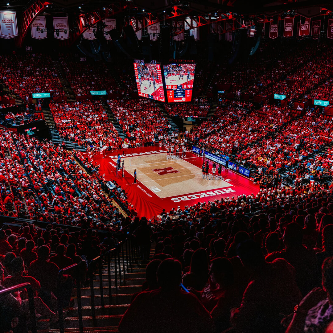 Packed arena with red-clad fans, volleyball players on court, and large digital screens overhead.