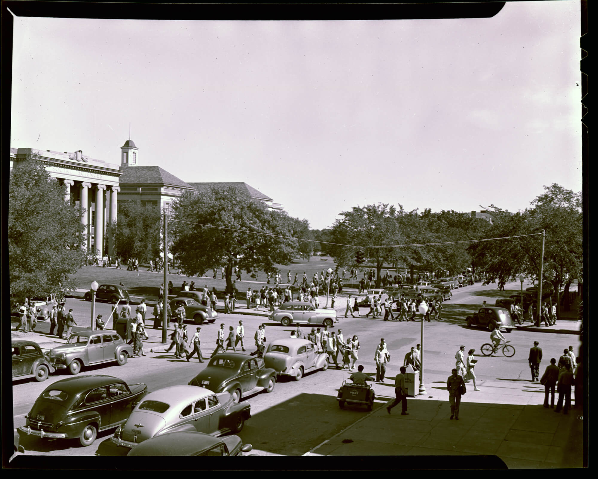 A black and white photograph of campus from the campus archives. The photo shows students walking across campus.