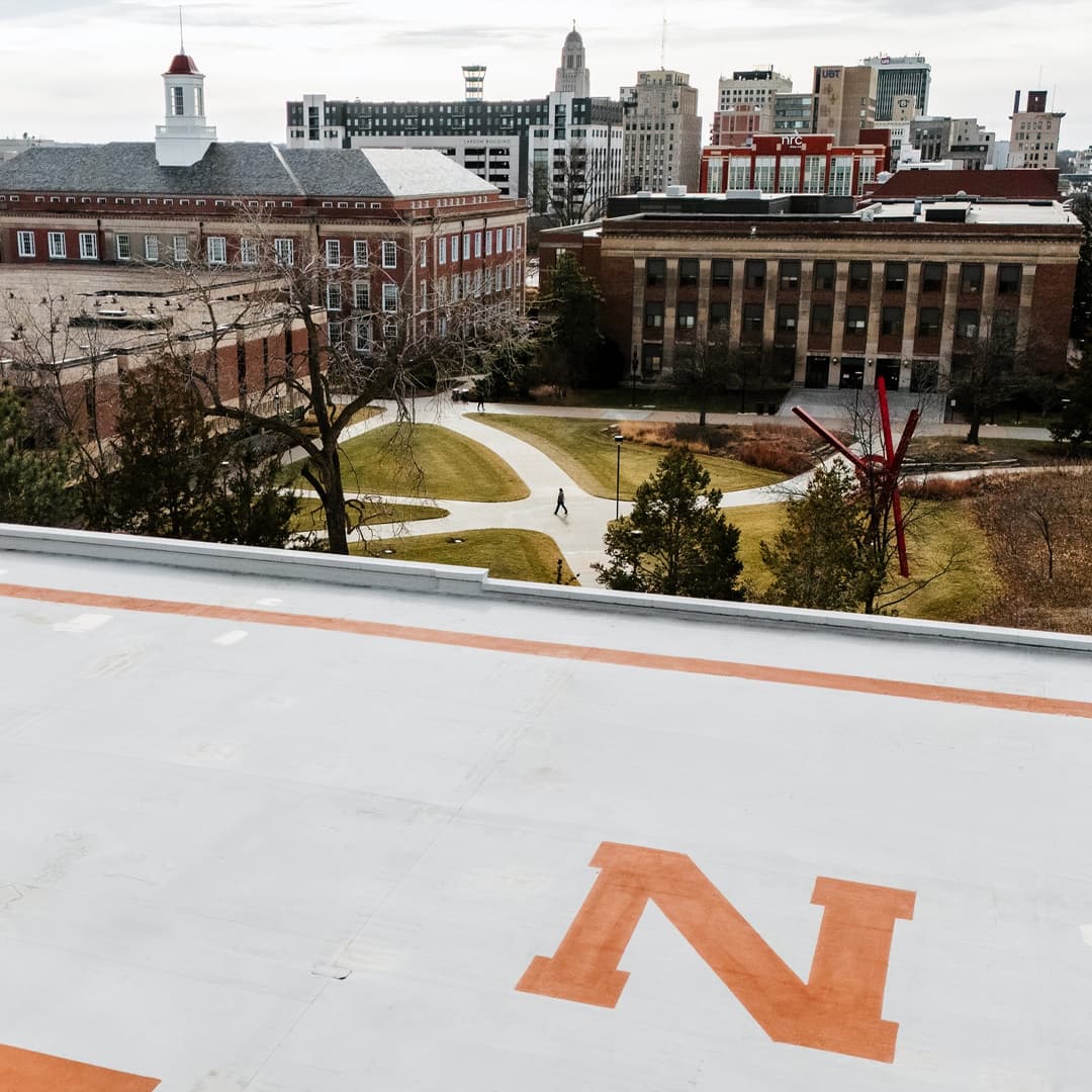 An aerial view of a Nebraska 'N' painted on the roof of a City Campus building. Campus sidewalks, buildings and the Lincoln skyline appear in the background.