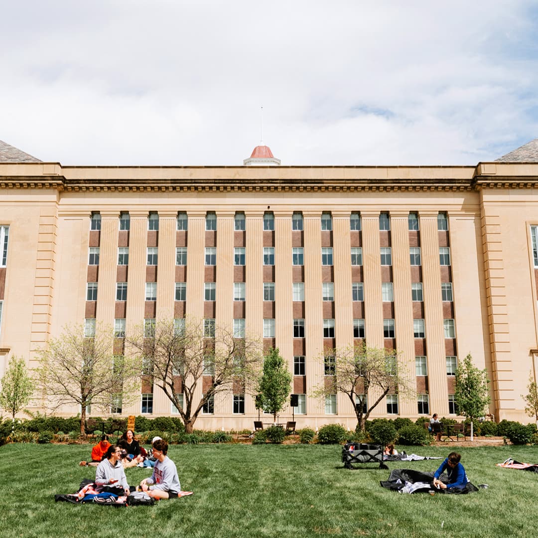 People sitting on a grassy lawn in front of Love Library under a cloudy sky.