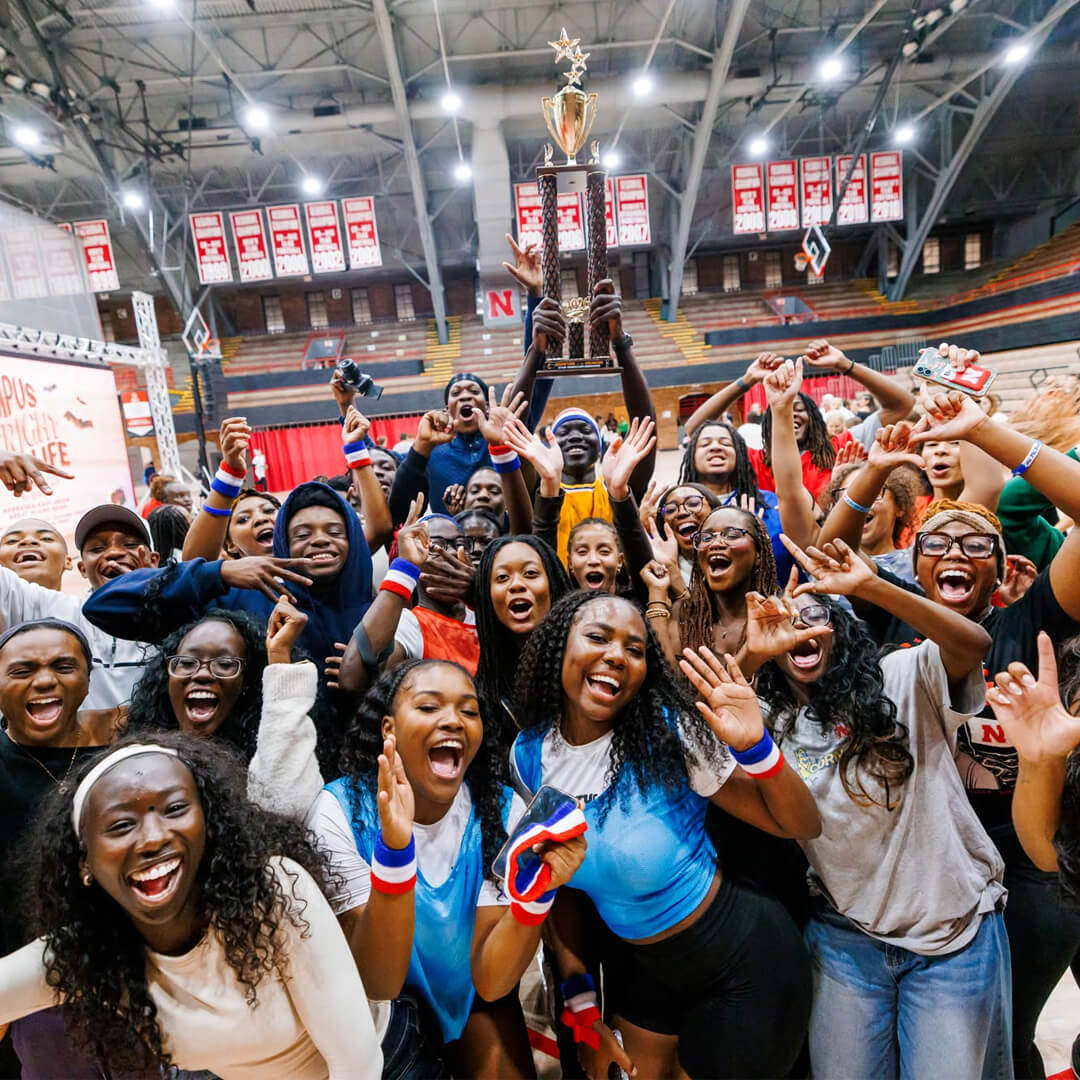 Group of students celebrating indoors under banners, holding a large trophy and cheering.