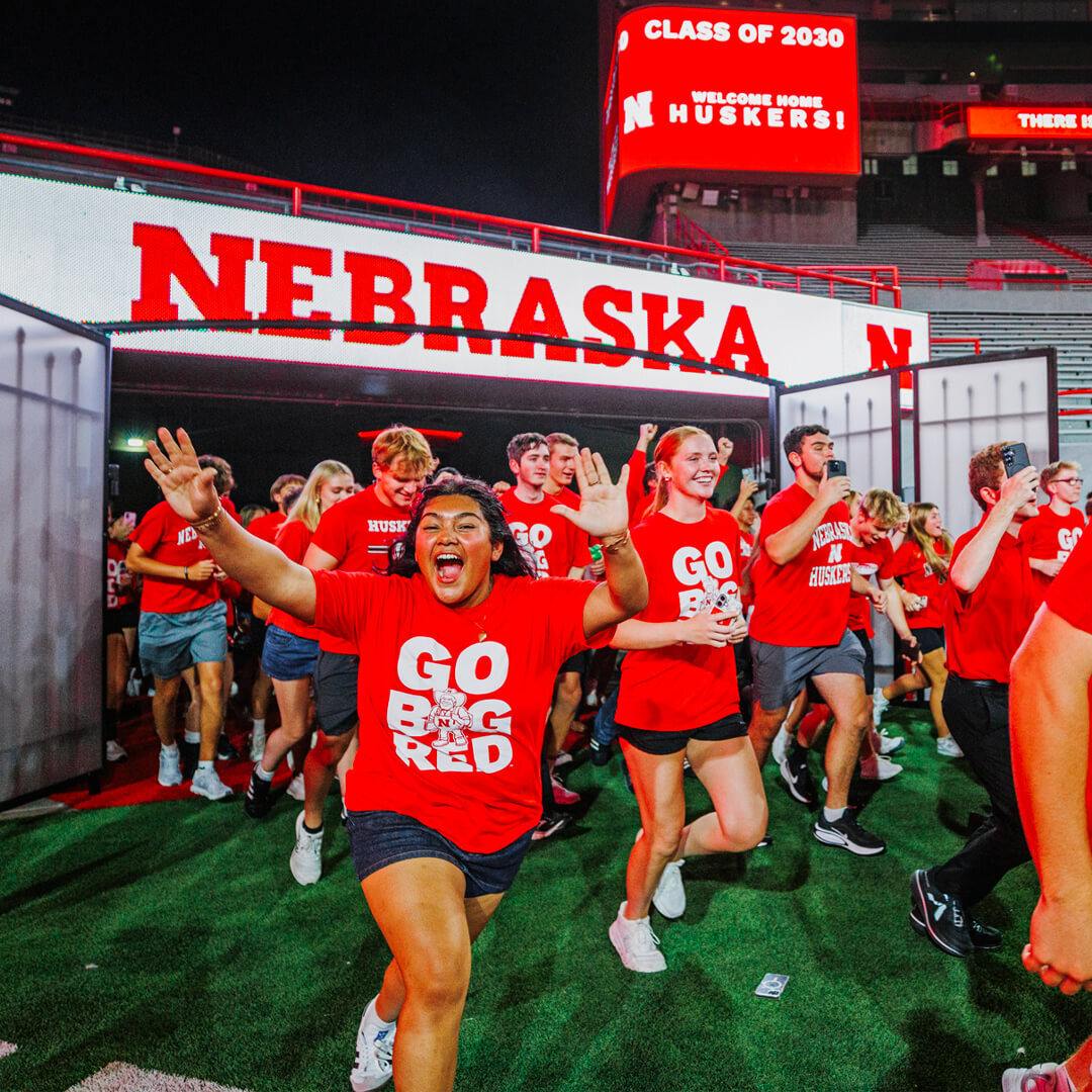 Students in red 'Go Big Red' shirts excitedly run into Memorial Stadium under a Nebraska sign for the Tunnel Walk.