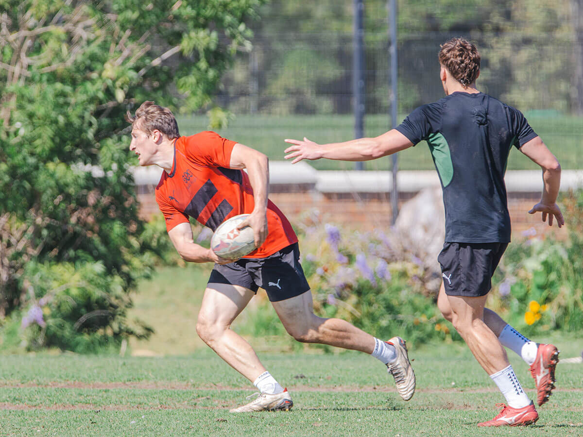 Rugby players on a grassy field, one with a ball evading the other's reach.