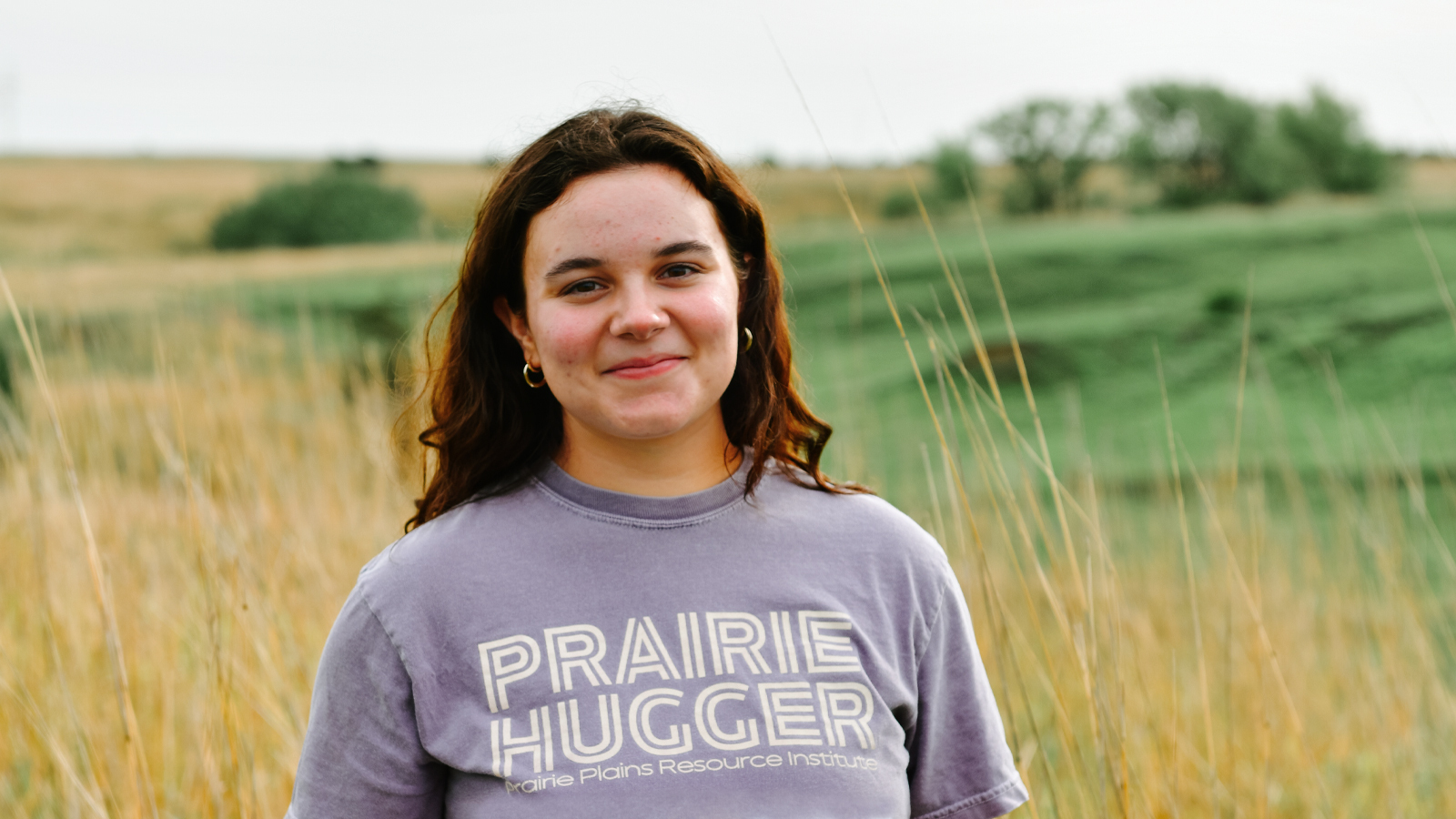 Person in a purple shirt standing in a grassy prairie with rolling green fields in the background.