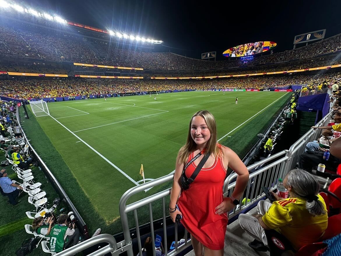 Elsa smiles in the stands at Kansas City Stadium. Behind her, the soccer field is brightly lit and fans fill the stadium for a night game.