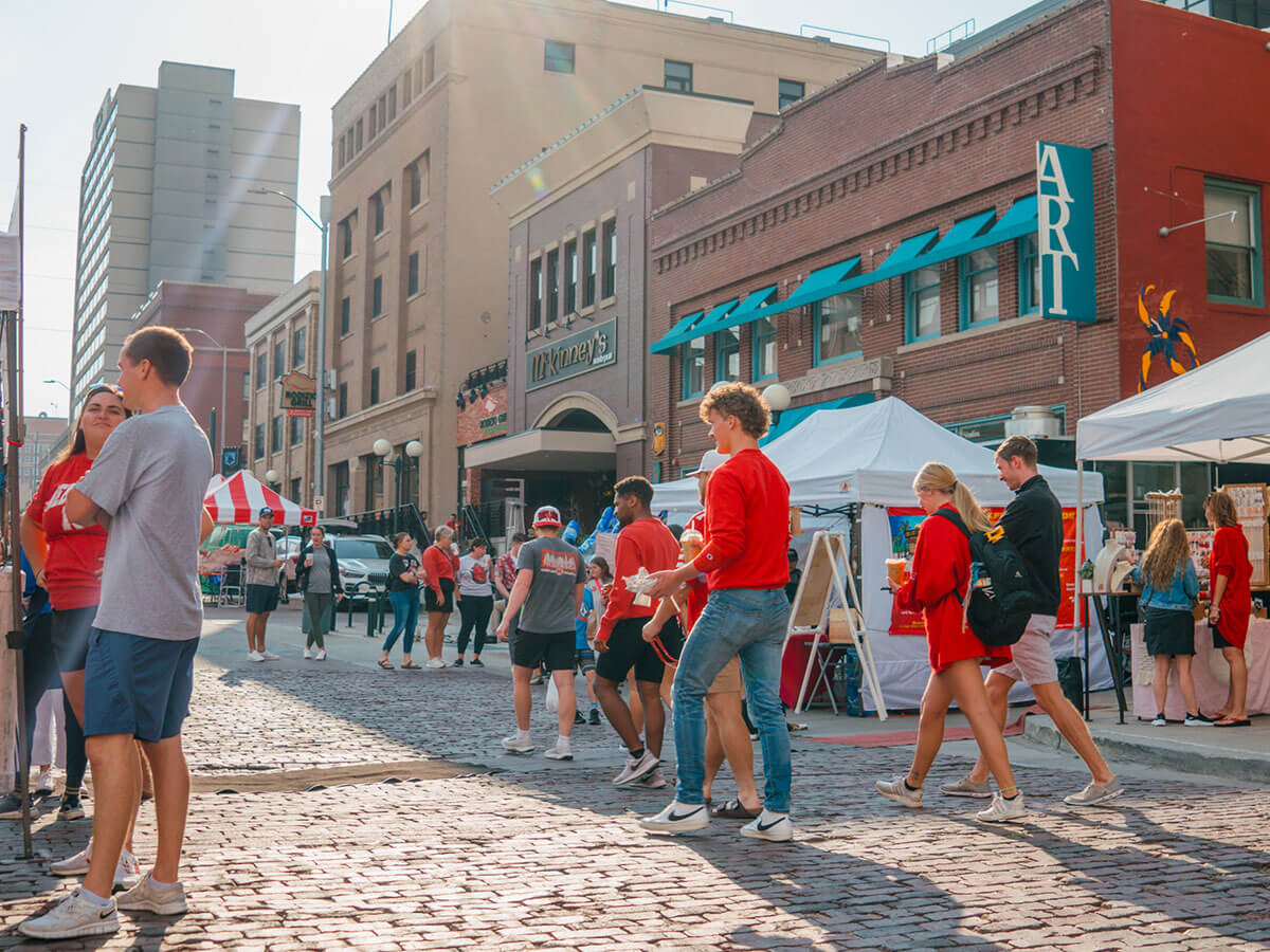 People stroll on a sunny cobblestone street with tents, shops, and a vibrant atmosphere during the Haymarket Farmers' Market..