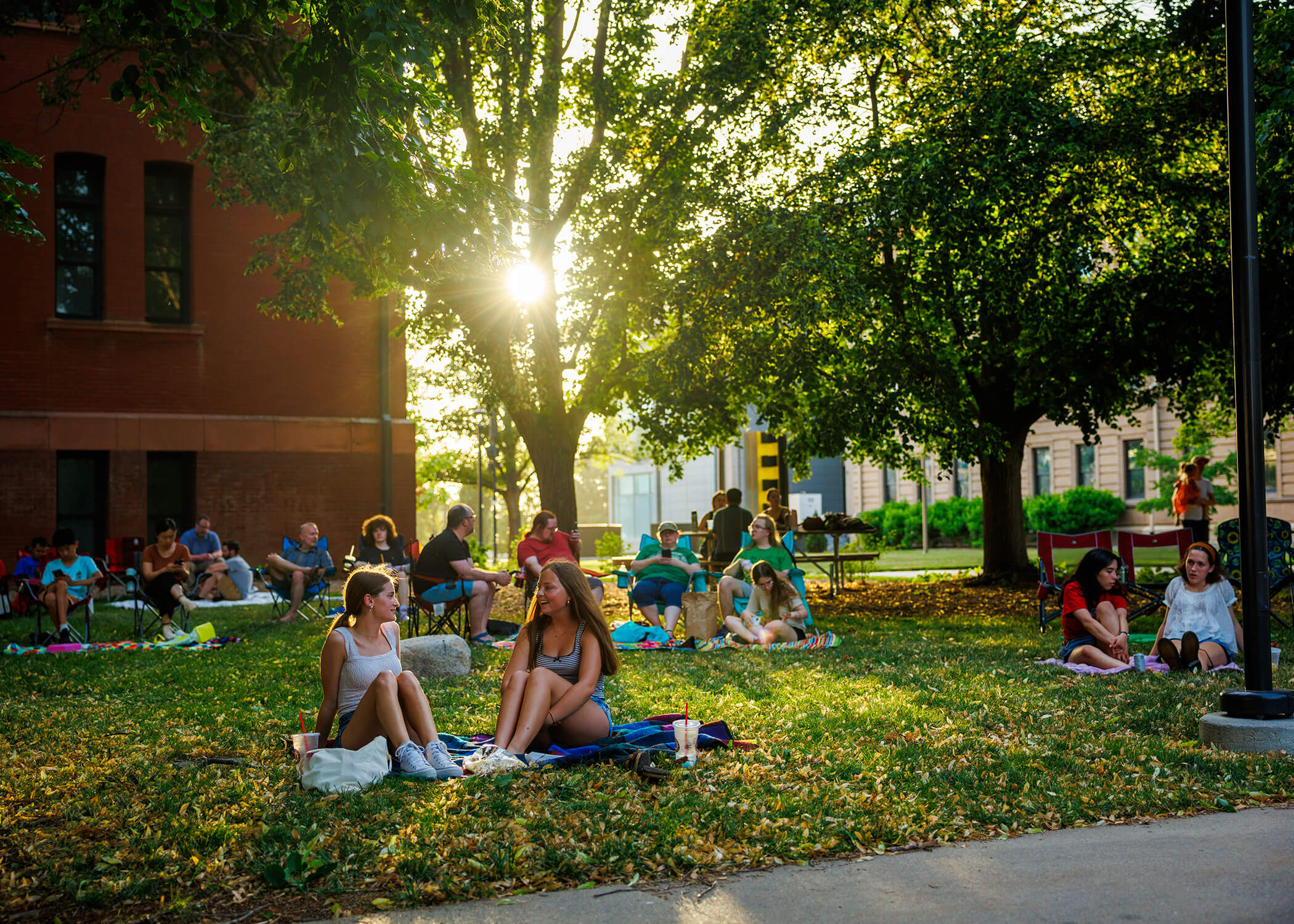 People relaxing on a sunny lawn, surrounded by trees and colorful blankets near a brick building during the Jazz in June series.