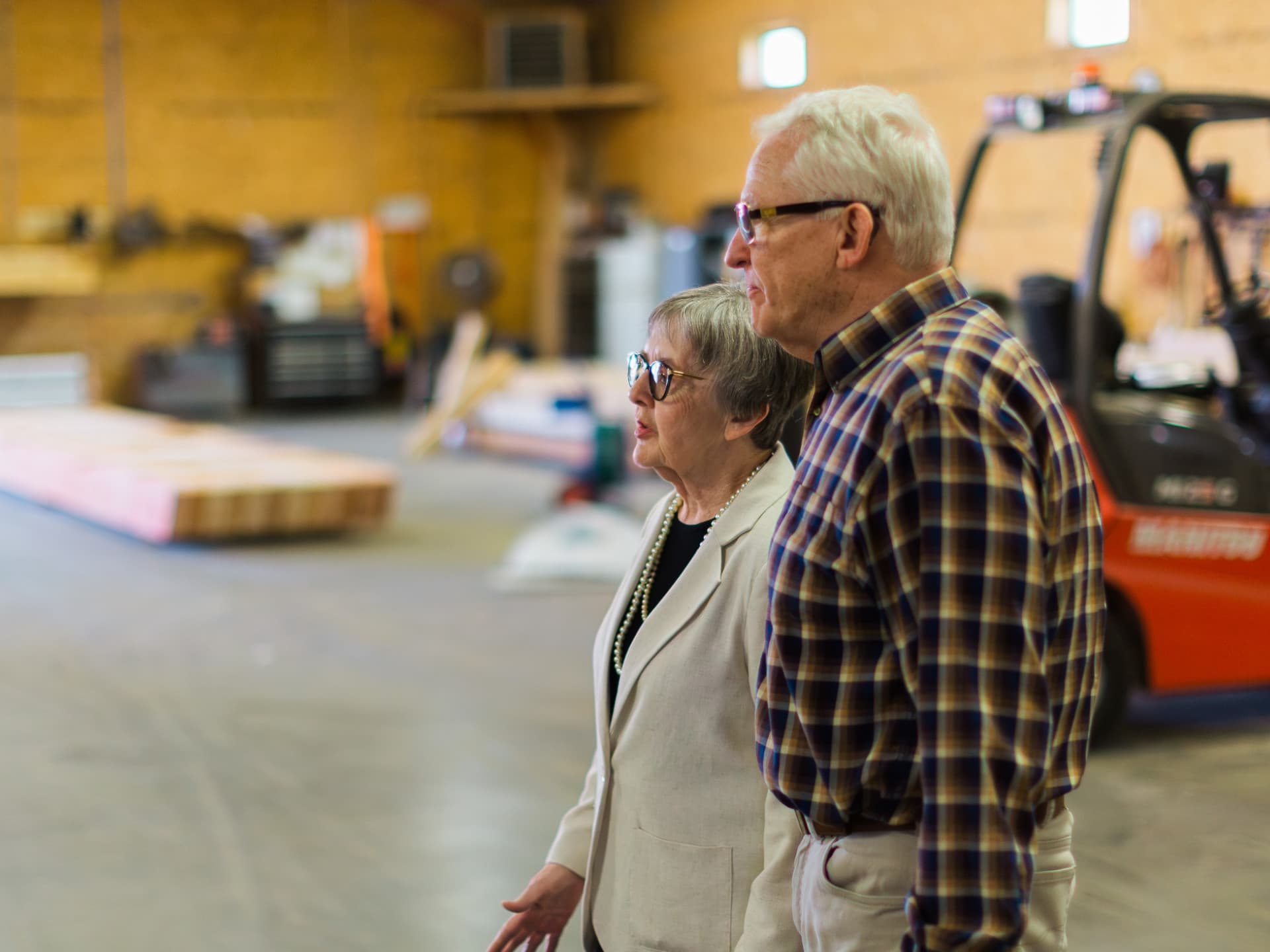 Elderly couple observing equipment in a warehouse with wooden walls and a forklift nearby.