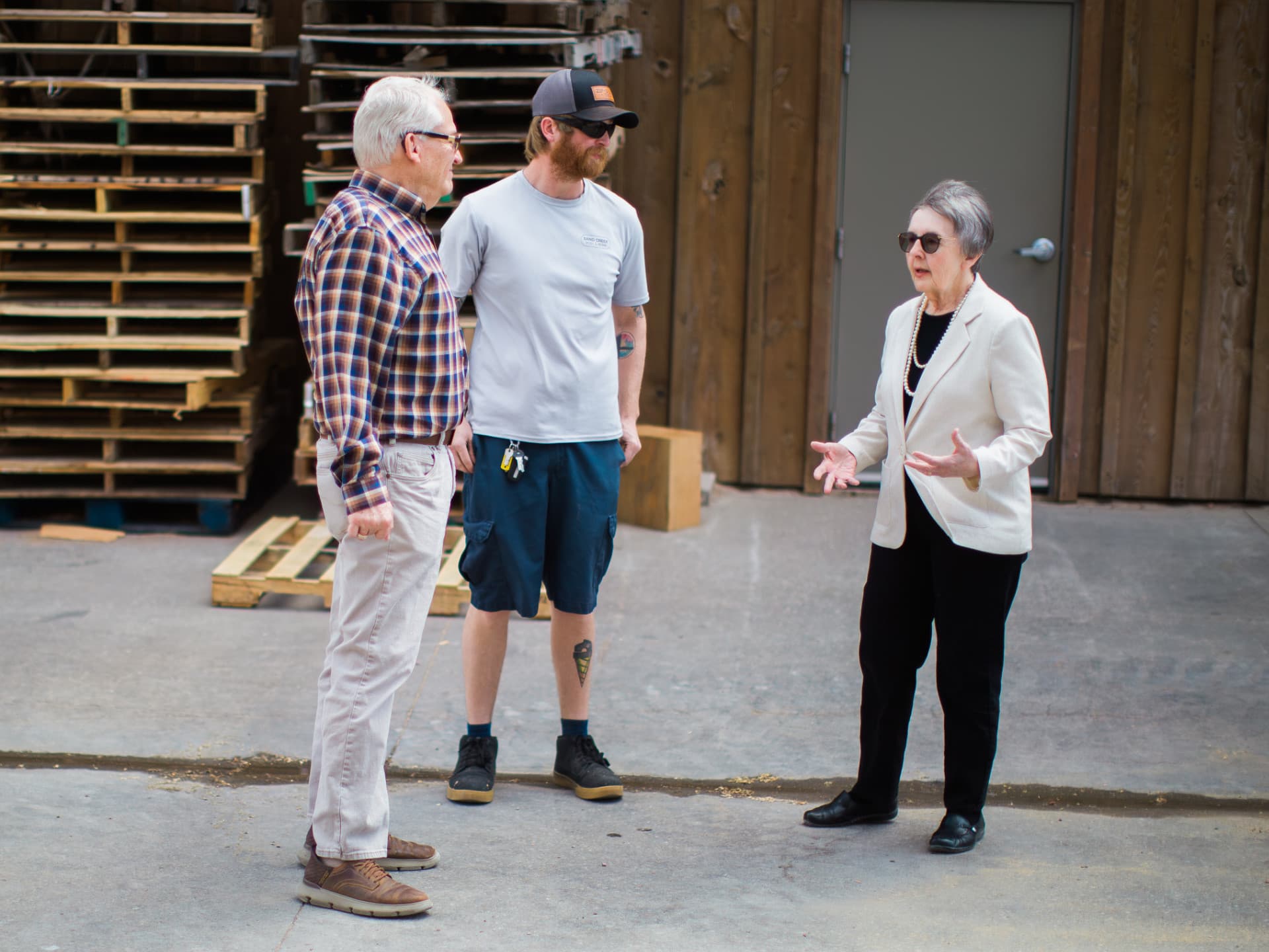 Three people converse outdoors near stacked wooden pallets.