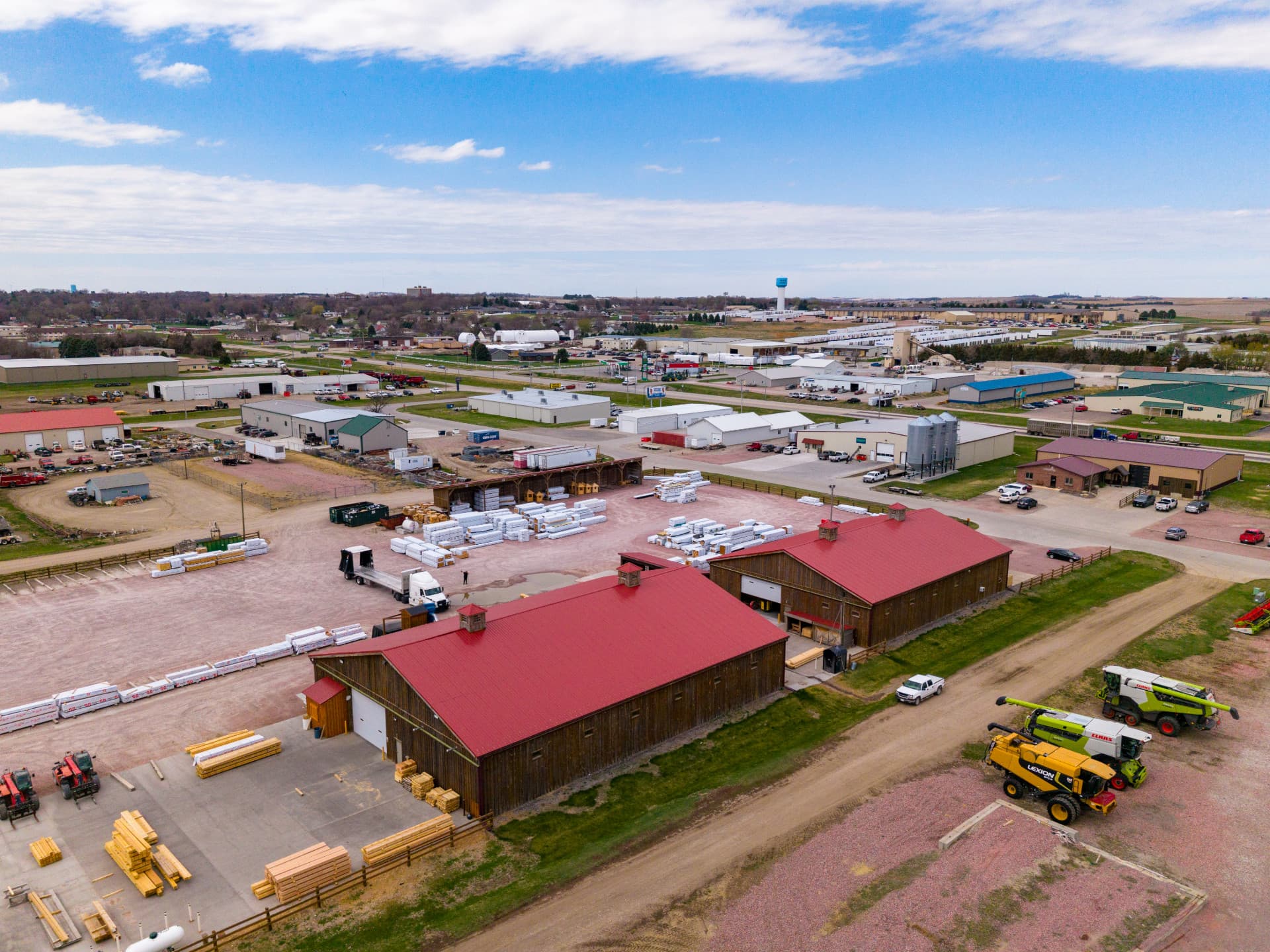 Aerial view of a rural lumberyard, warehouses with red roofs, and farm equipment nearby.