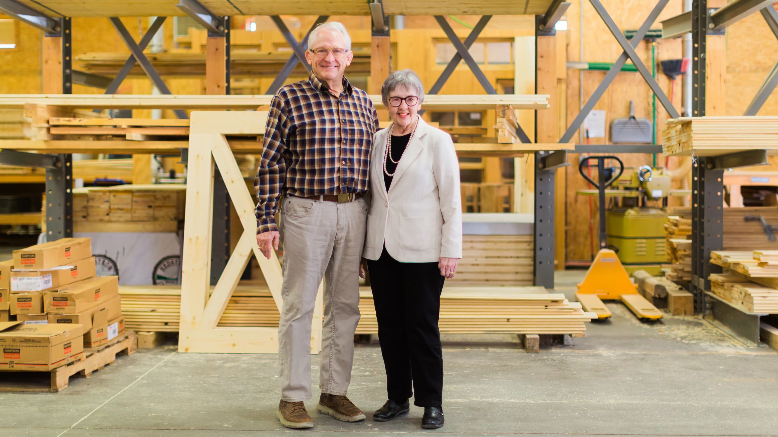 Man and woman standing in a wood workshop, surrounded by lumber and tools, smiling warmly.