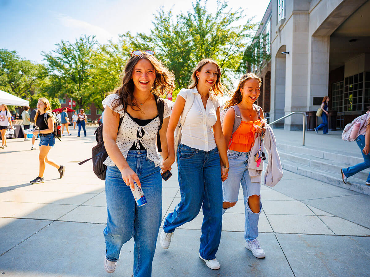 Three women smiling and walking outdoors on a sunny day, surrounded by trees and buildings.