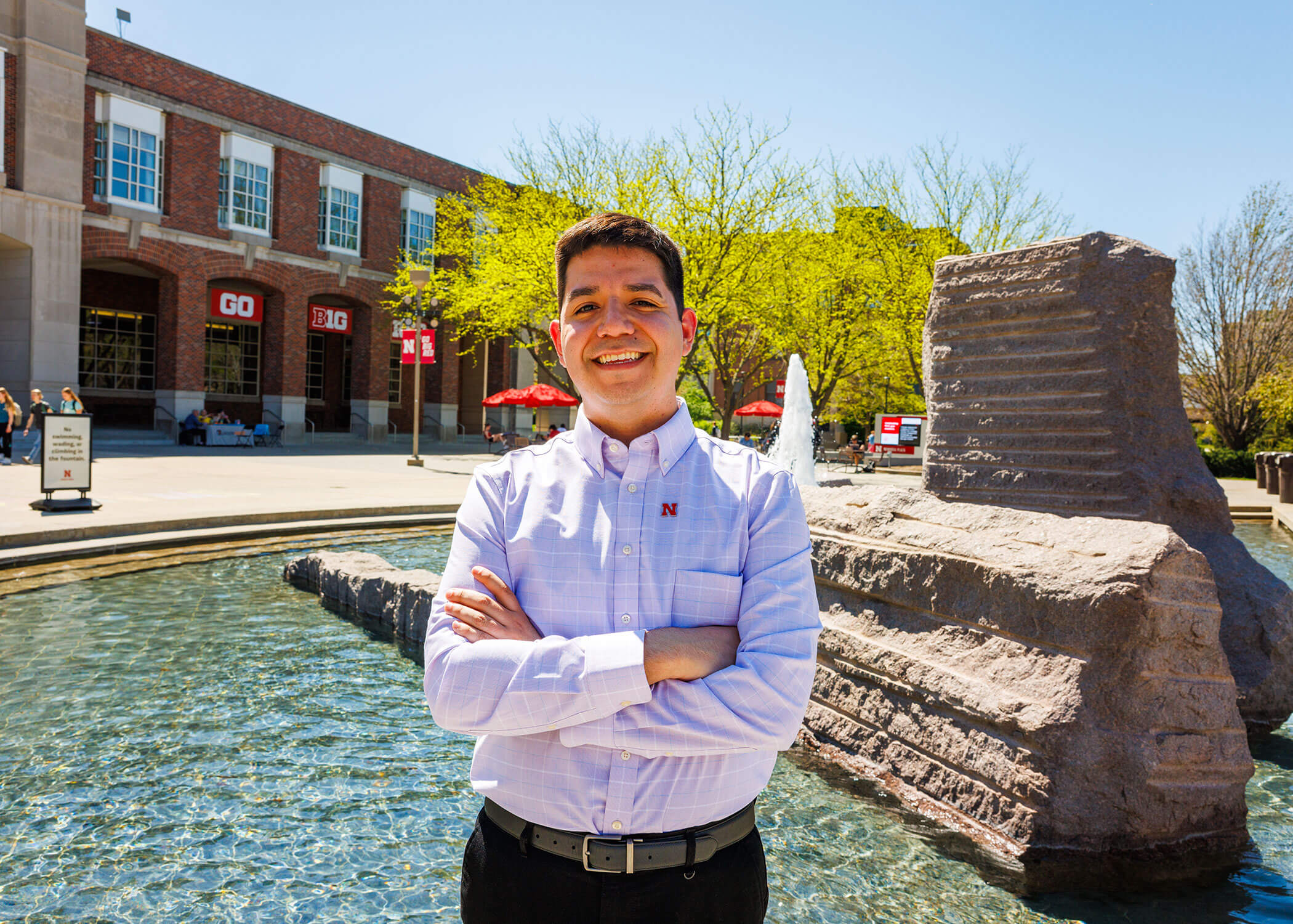 Lupe Perez standing by fountain, arms crossed on sunny day with the Nebraska Union set in the background.