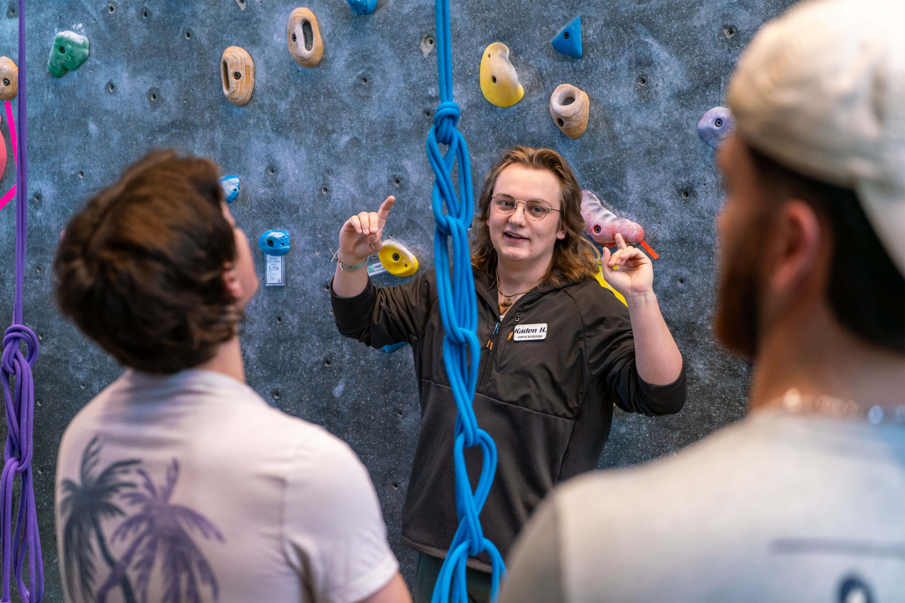 Student standing in front of climbing wall gesturing to other students